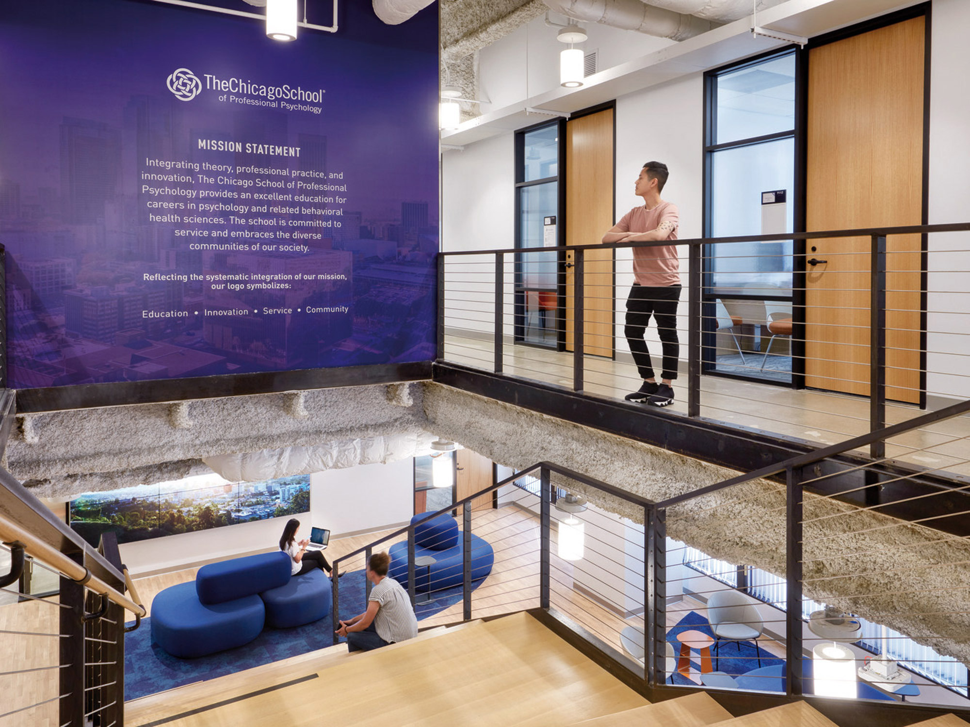 Open-plan office space with a multi-level layout featuring natural wood finishes and glass partitions. Blue curved sofas complement the informal seating area, with a woman standing at the balcony overlooking the workspace. The Chicago School mission statement adorns the wall, enhancing the educational environment.