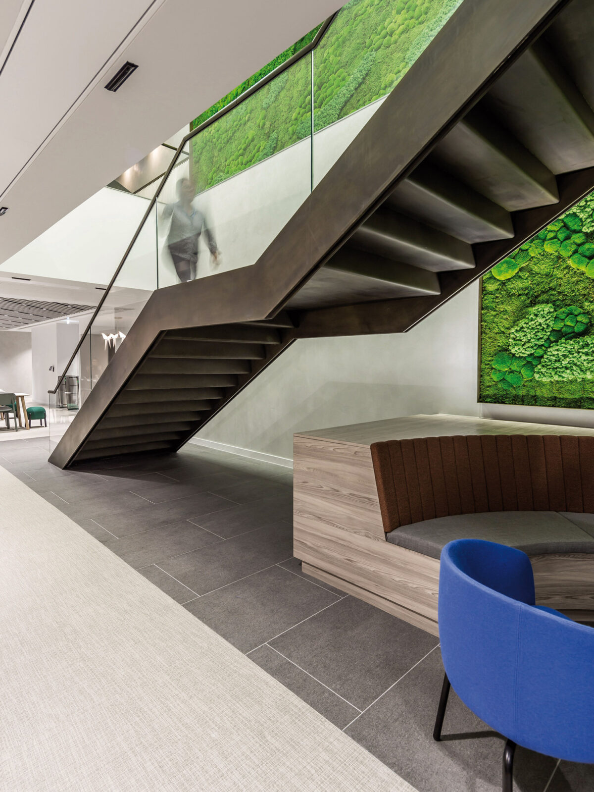 Sleek modern office space featuring a floating staircase with glass balustrades adjacent to a vibrant green living wall. Wooden bench seating and a pop of color from a blue armchair create a welcoming break area.