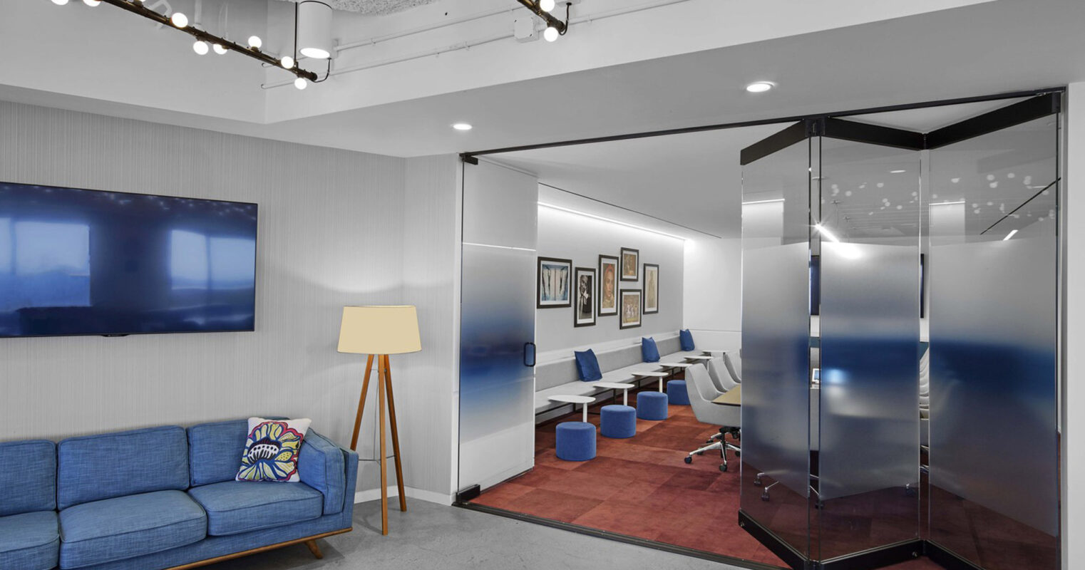 Modern office lounge with a vibrant blue upholstered couch, complemented by a minimalist floor lamp. Track lighting illuminates the space, leading to a glass-enclosed meeting area with sleek chairs and mounted digital displays.