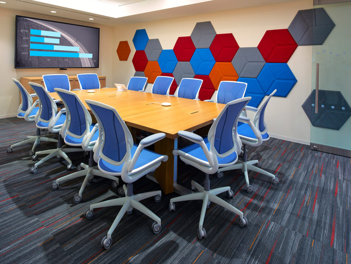 Modern conference room featuring a large, rectangular wooden table with blue ergonomic chairs, vibrant geometric acoustic panels on the wall, and a patterned gray carpet with red accents.