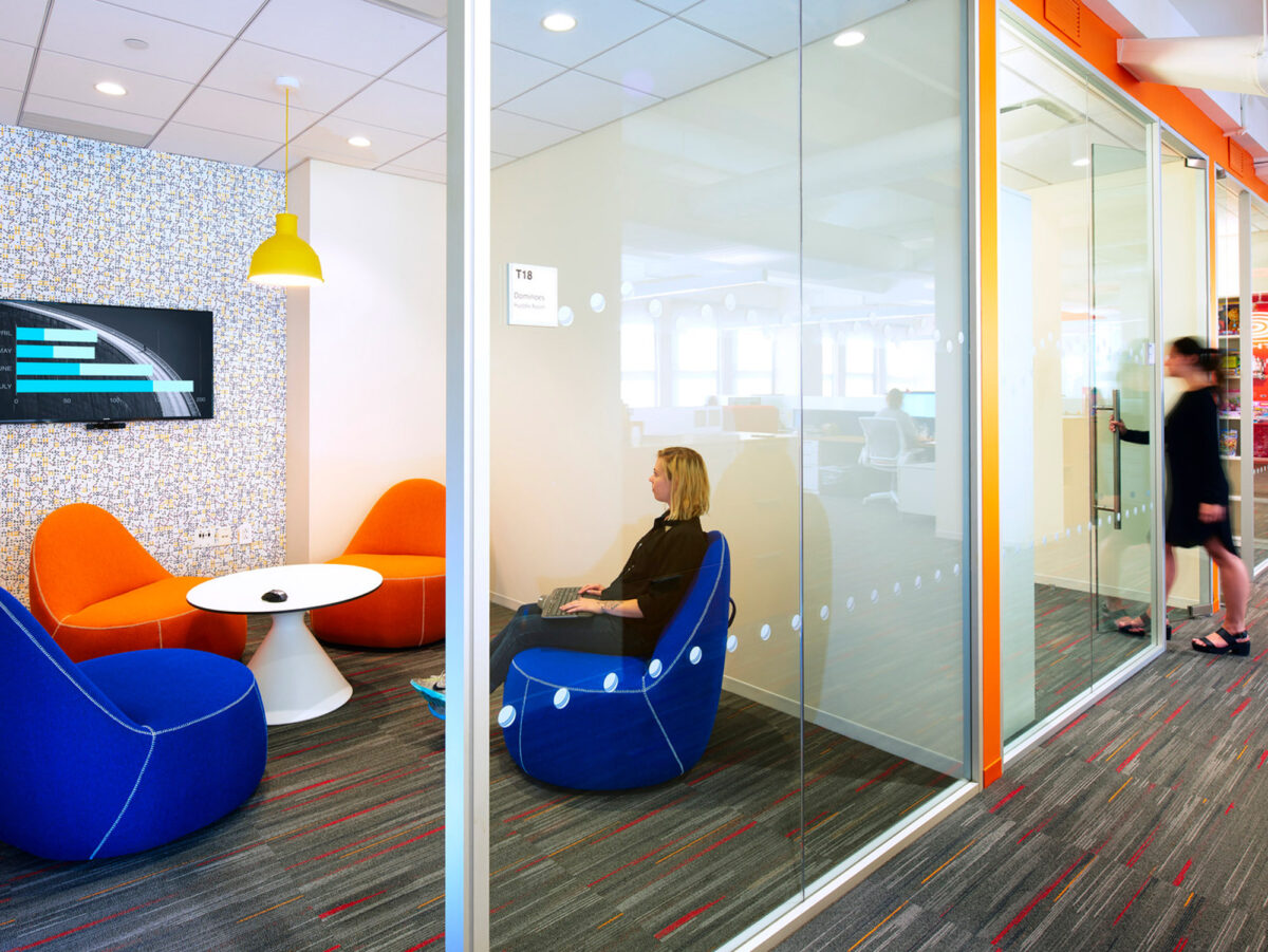 Bright, modern office breakout area featuring bold orange chairs with white accents, a mosaic-tiled wall, and neutral carpet with vibrant colored streaks. Glass walls maintain a transparent, open feel while a yellow pendant light adds a pop of contrasting color.