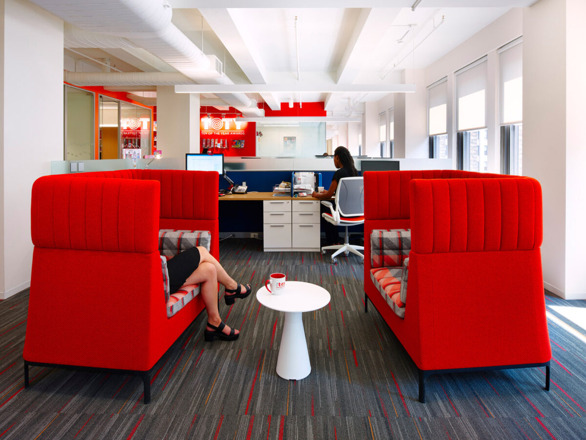 Modern office lounge with vibrant red high-backed sofas, white circular coffee table, and a woman seated working on a laptop. The space features bright natural light, contrasting gray carpet with colorful accents, and glass partition walls.