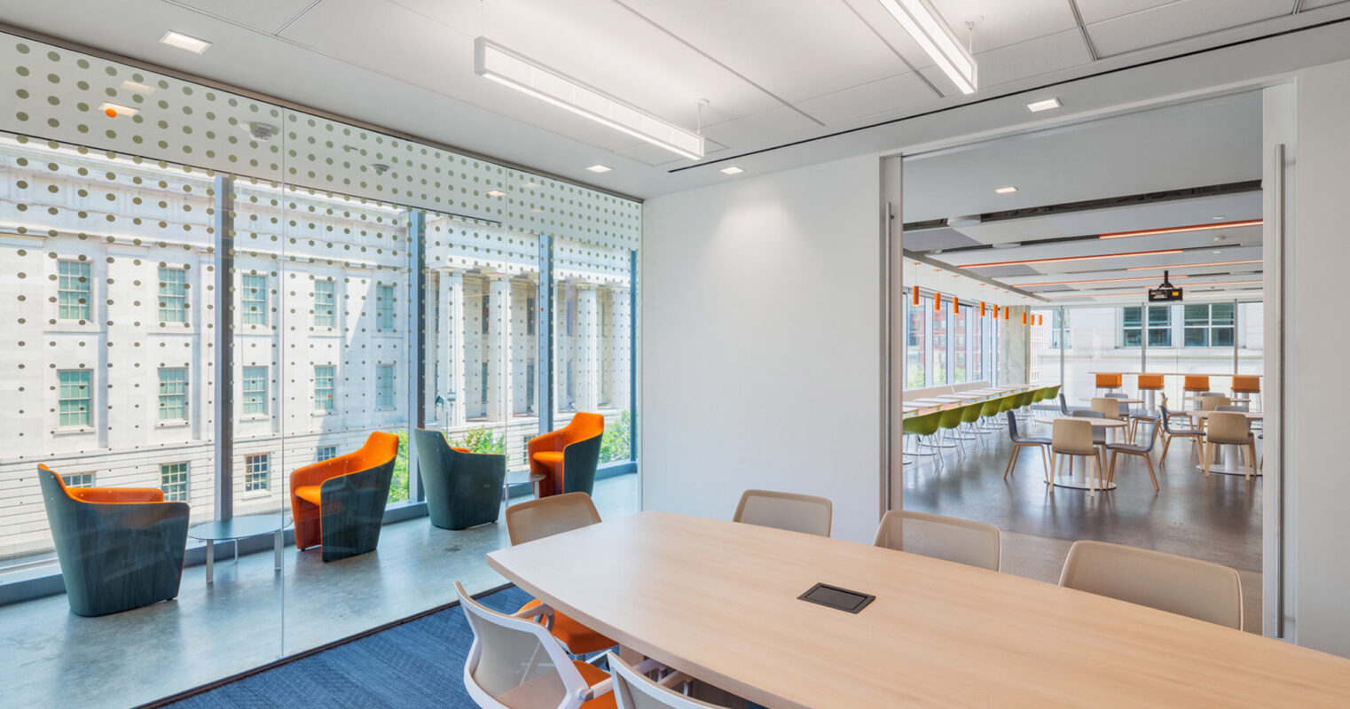 Modern open-concept office space with natural light filtering through patterned window screens. Sleek, light wood conference table paired with orange and white chairs, complemented by a casual dining area in the background with pendant lighting. Vibrant orange accents energize the room.