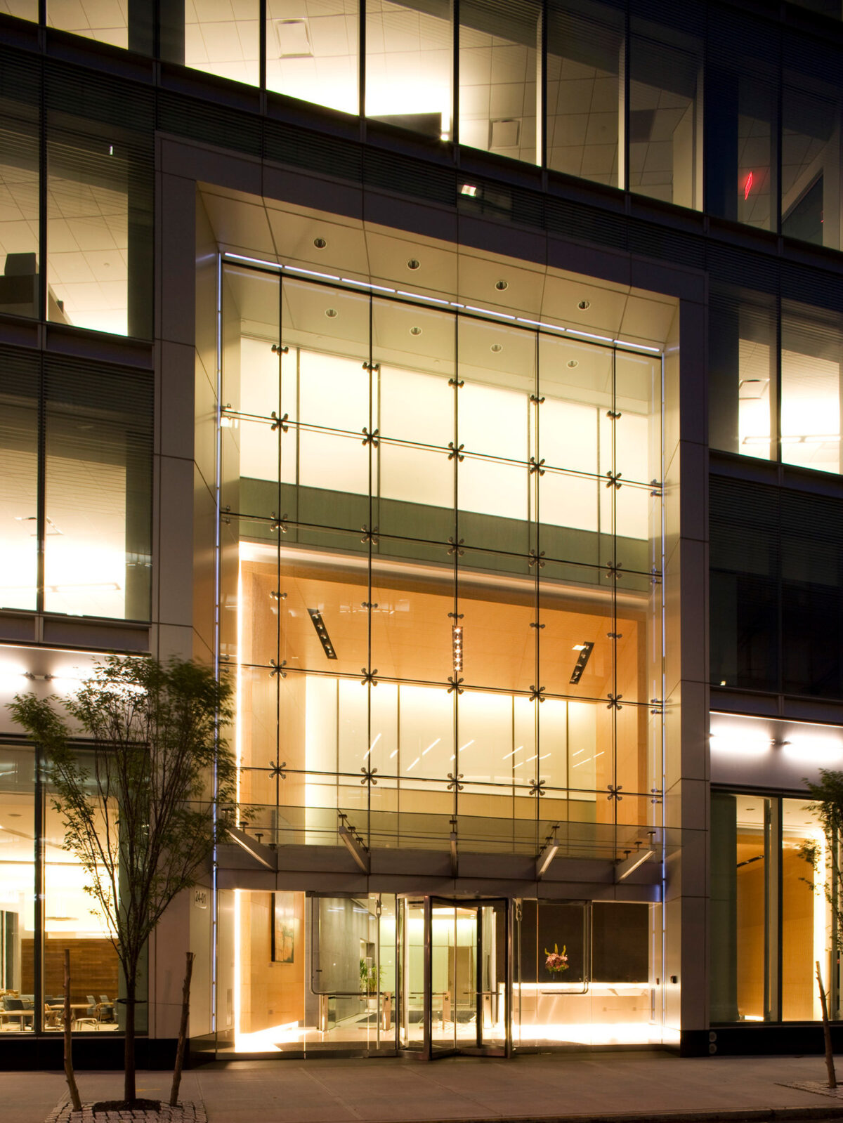 Modern office building entrance featuring a tall glass facade with a grid of metallic mullions, illuminated by warm interior lighting against the evening sky, showcasing transparency and contemporary design aesthetics.