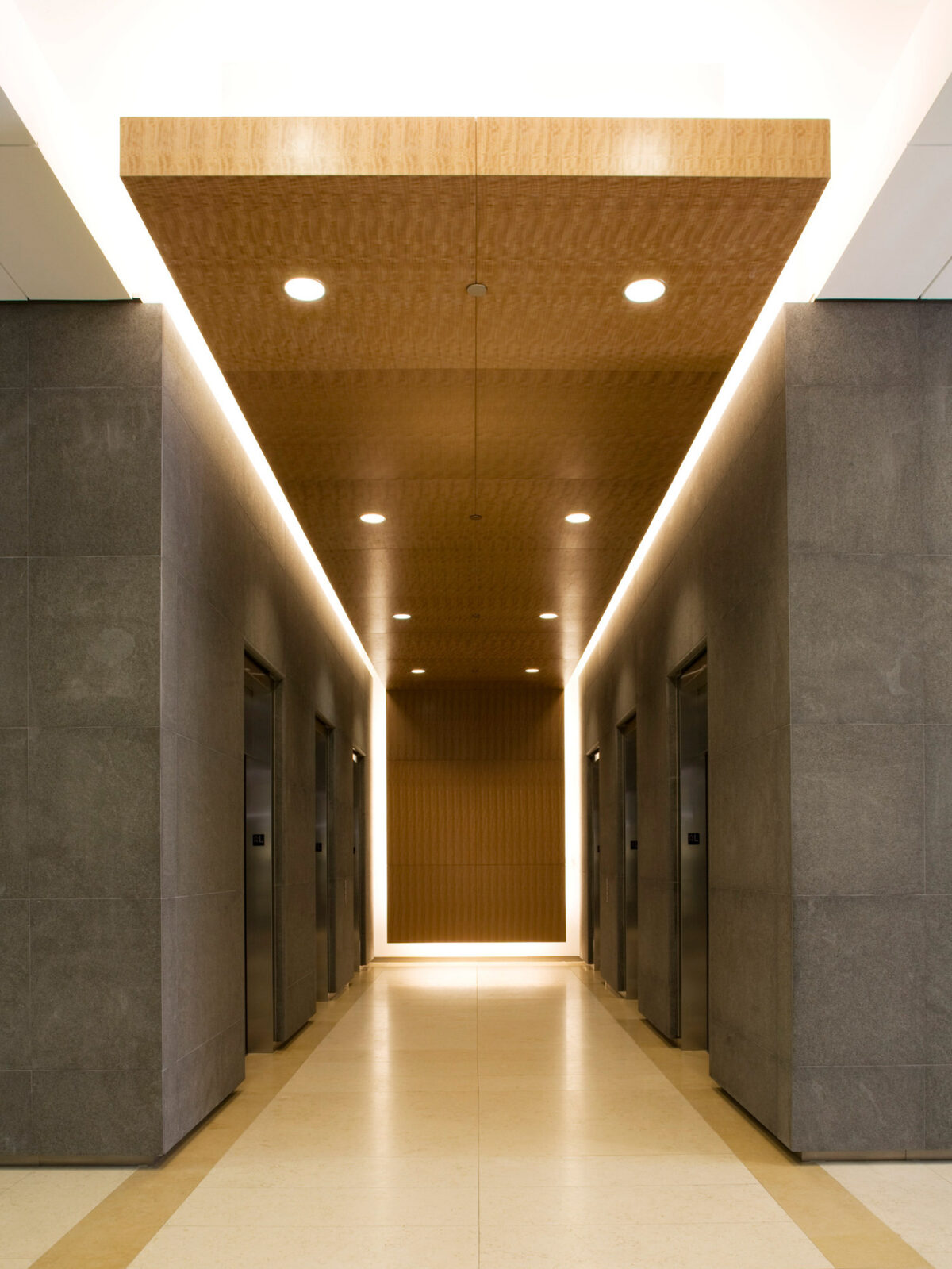 Modern hallway with recessed lighting emphasizing the straight lines and planes. Neutral tones of the walls contrast with a warm wooden ceiling and floors, highlighting minimalist aesthetic and architectural symmetry.