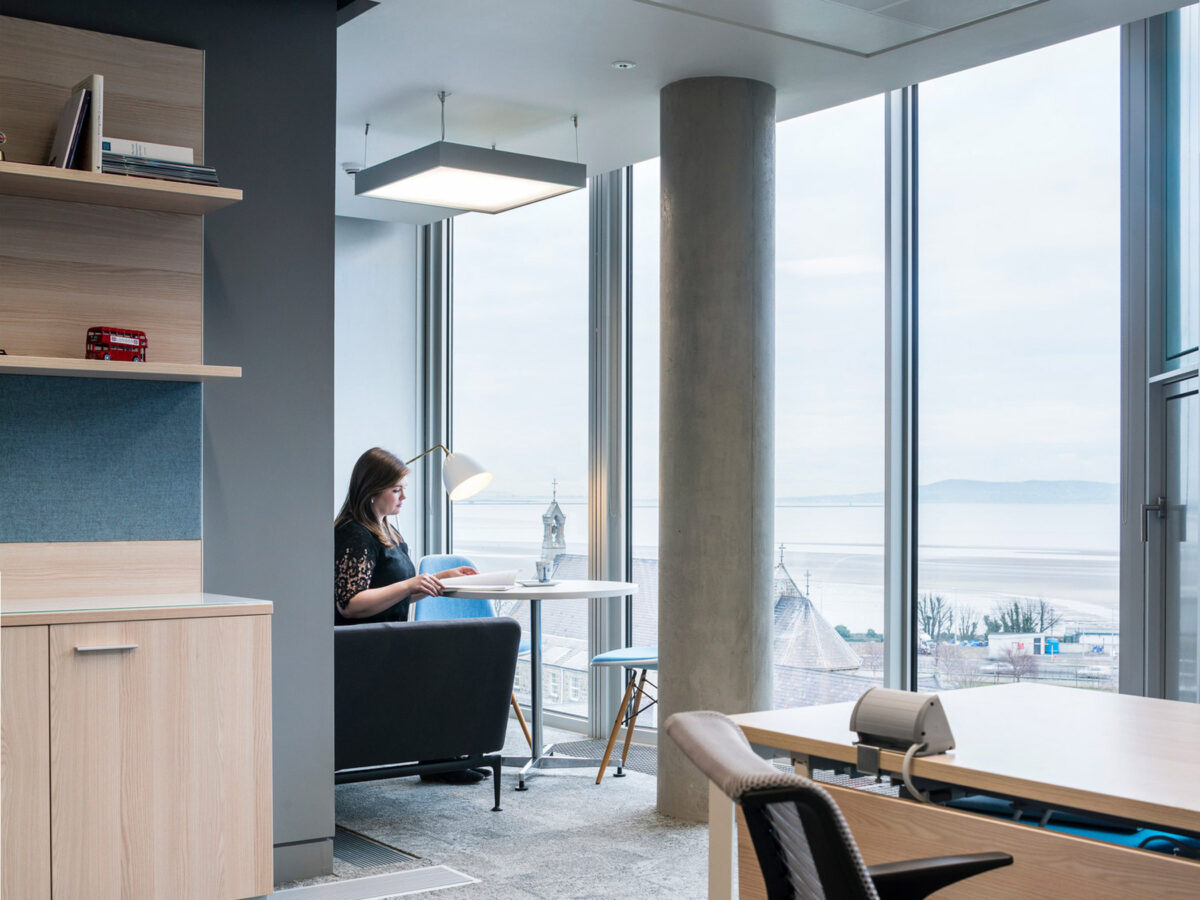 Modern home office with minimalist design, featuring a light wood desk, black chair, and built-in wall shelving. A woman sits reading near a floor-to-ceiling window overlooking a calm water view, illuminated by natural light and a sleek hanging pendant lamp.