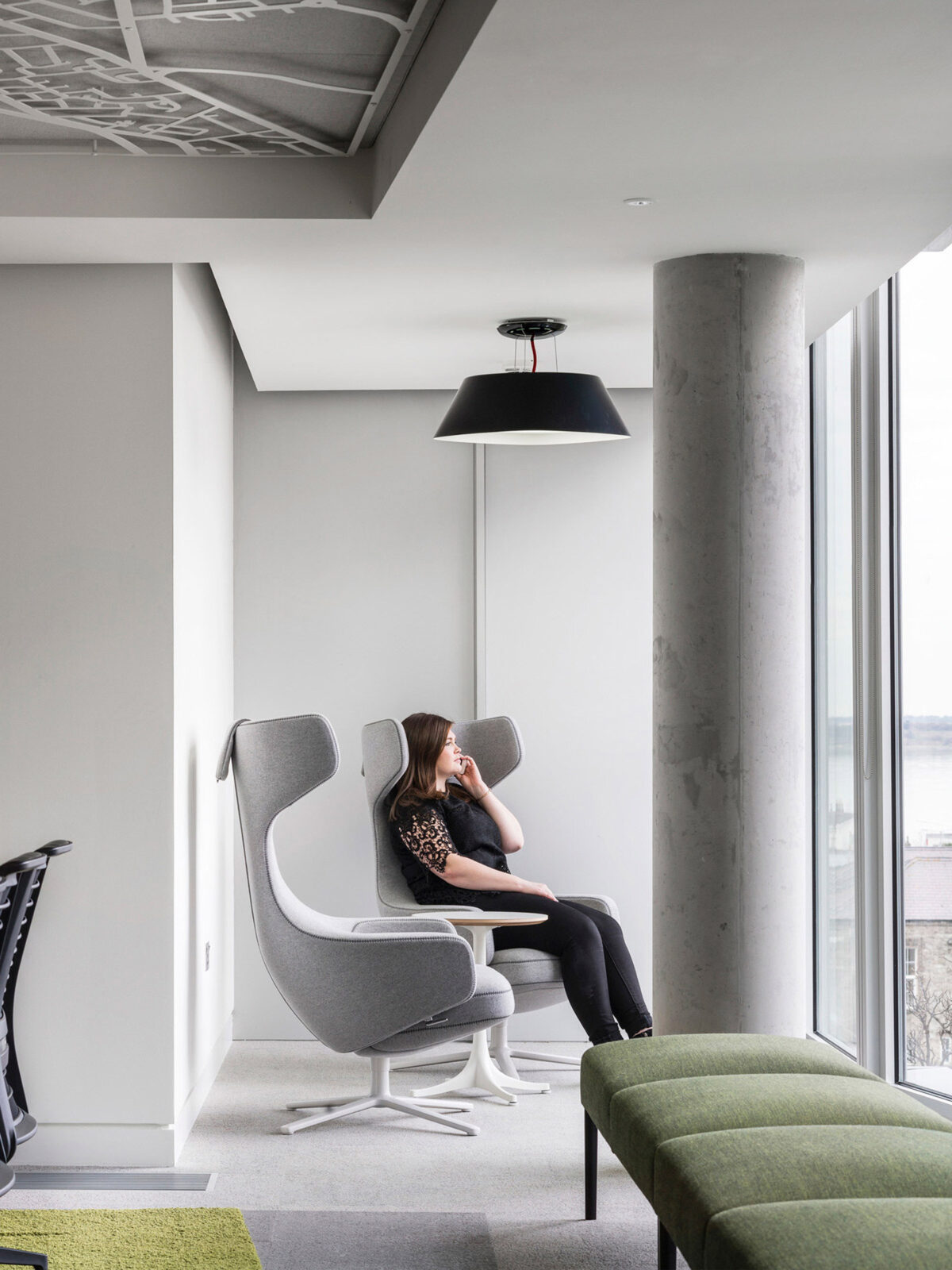 Modern interior with an exposed concrete column, floor-to-ceiling windows, and an open ceiling revealing ductwork. A woman lounges in a gray, wingback swivel chair, complementing a green linear sofa, against the backdrop of urban views, marrying industrial and cozy design elements.