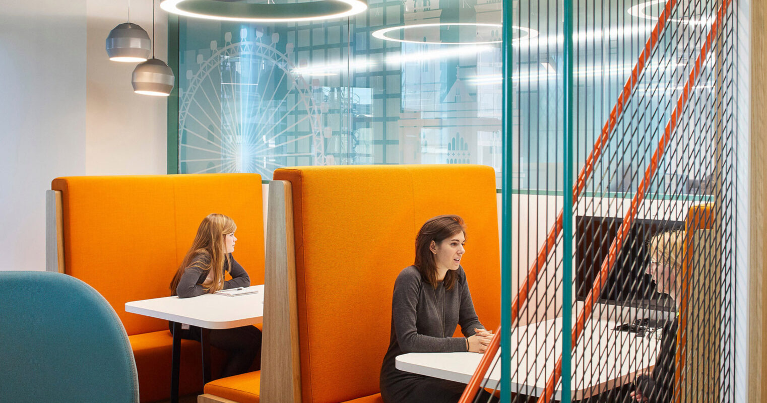 Modern office breakout space with vibrant orange privacy booths, complemented by teal chairs. Overhead, industrial-style pendant lights illuminate the area. Behind a partition featuring vertical colored cords, a collaborative workspace with a staircase is subtly visible.