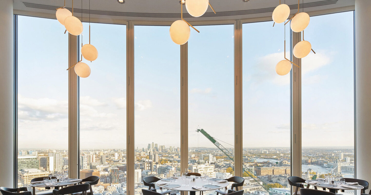 Elegant dining space featuring floor-to-ceiling windows providing panoramic city views, complemented by warm wooden flooring and spherical pendant lights. Scandinavian-inspired chairs and simple, chic tables set a minimalist yet inviting tone.