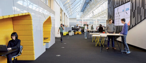 Modern office interior with an open floor plan, featuring angular lighting fixtures overhead and a vibrant yellow seating nook. Collaborative spaces with high-top tables encourage interaction, while the far wall showcases project displays under a naturally lit atrium.