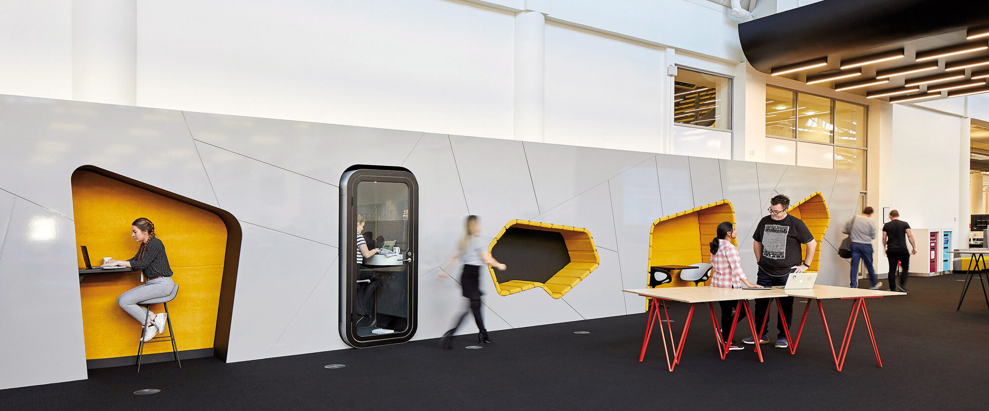 Modern office interior featuring honeycomb-inspired work pods with vibrant yellow accents, providing private nooks for concentration. The open floor plan includes high ceilings and skylights, enhancing natural light and spaciousness. A communal work table with red stools encourages collaboration.