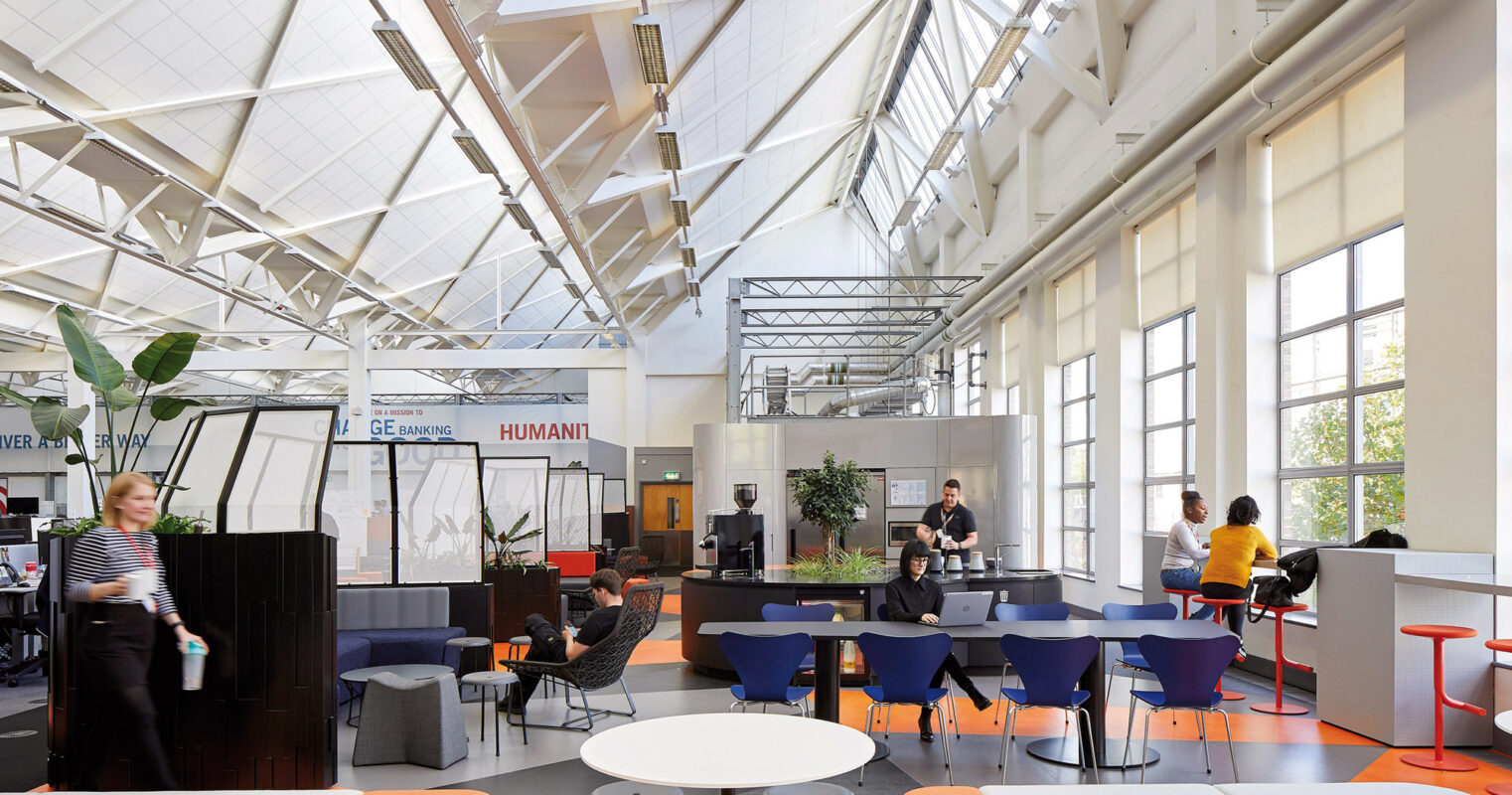 Bright, open-plan office space with high white ceilings and skylights that allow natural light to flood the interior. The layout features a mix of casual seating areas with vibrant orange and blue chairs, communal worktables, and private, black-paneled work pods, promoting a flexible and modern work environment.