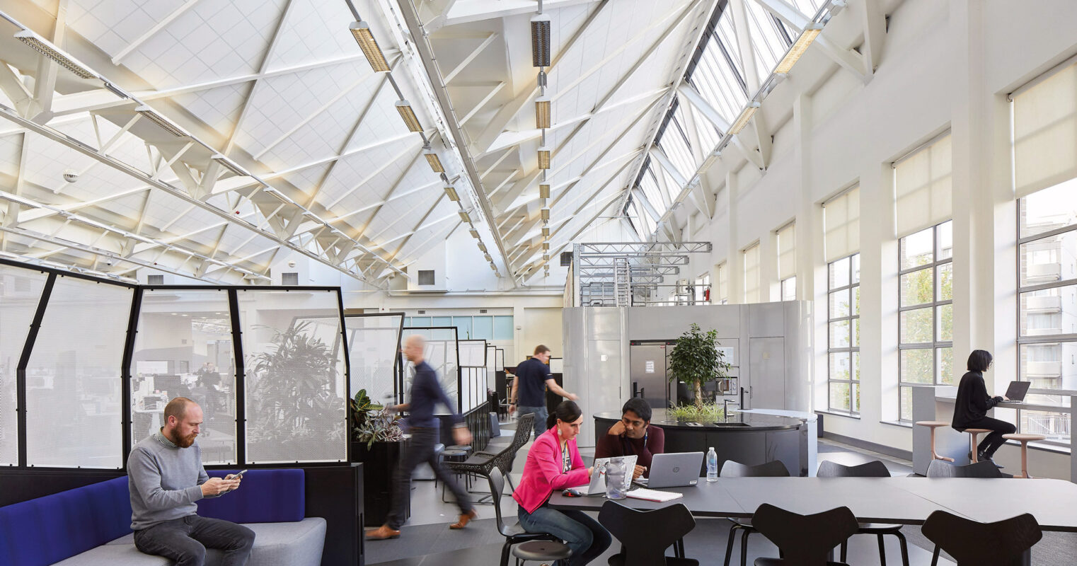 Modern office space featuring a high vaulted ceiling with skylights, exposed white trusses, and clerestory windows, ensuring abundant natural light. Varied workstations with ergonomic furniture are arranged in an open-plan layout, encouraging collaboration. Transparent partition walls provide privacy without sacrificing openness or light flow.