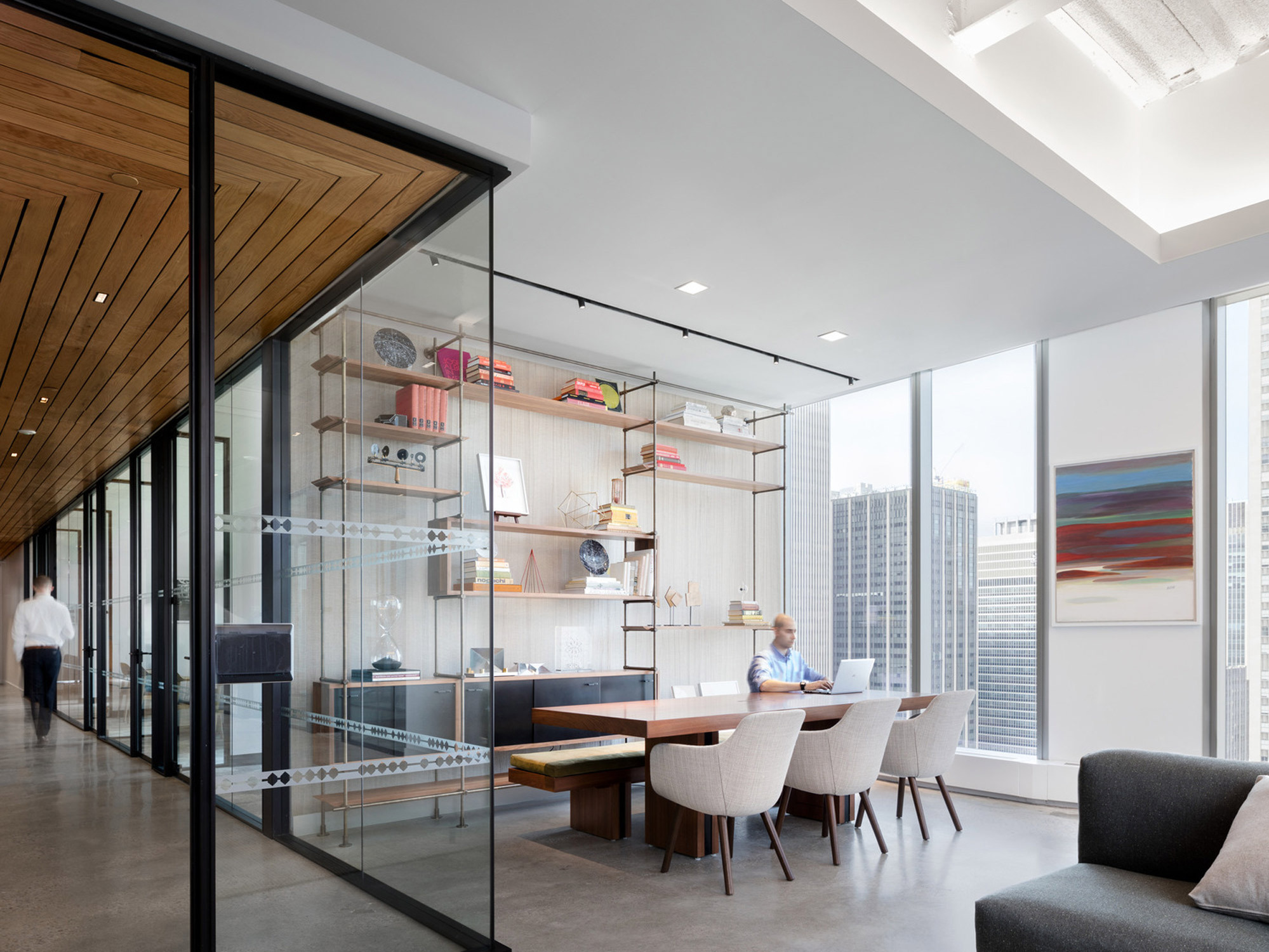 Open-plan office space with floor-to-ceiling windows offering abundant natural light, minimalist furniture, and a warm wood slat ceiling. An elegant glass partition separates the meeting area adorned with a shelving unit showcasing decorative objects.