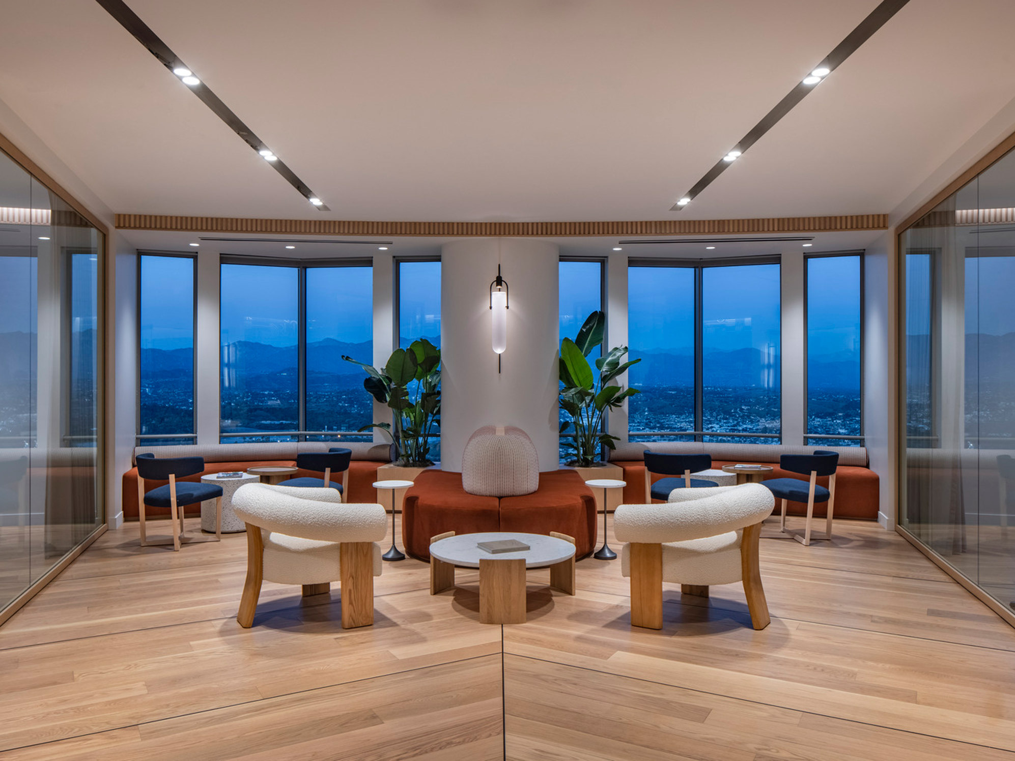 Modern interior showcasing a symmetrical layout with natural wood floors, recessed lighting, and floor-to-ceiling windows overlooking a cityscape. Central conversation area features contemporary furniture with organic lines, accented by potted plants and a statement pendant light.