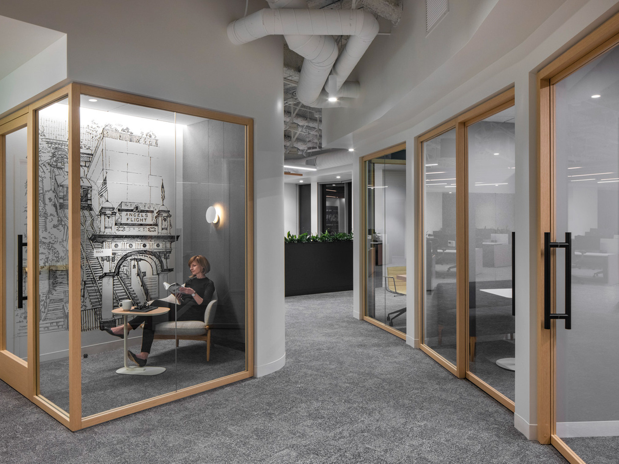 Modern office nook featuring glass walls, wooden door frames, and a monochromatic color scheme. A person sits at a minimalist desk inside, adjacent to a large-scale wall mural, under exposed ceiling infrastructure and soft pendant lighting.