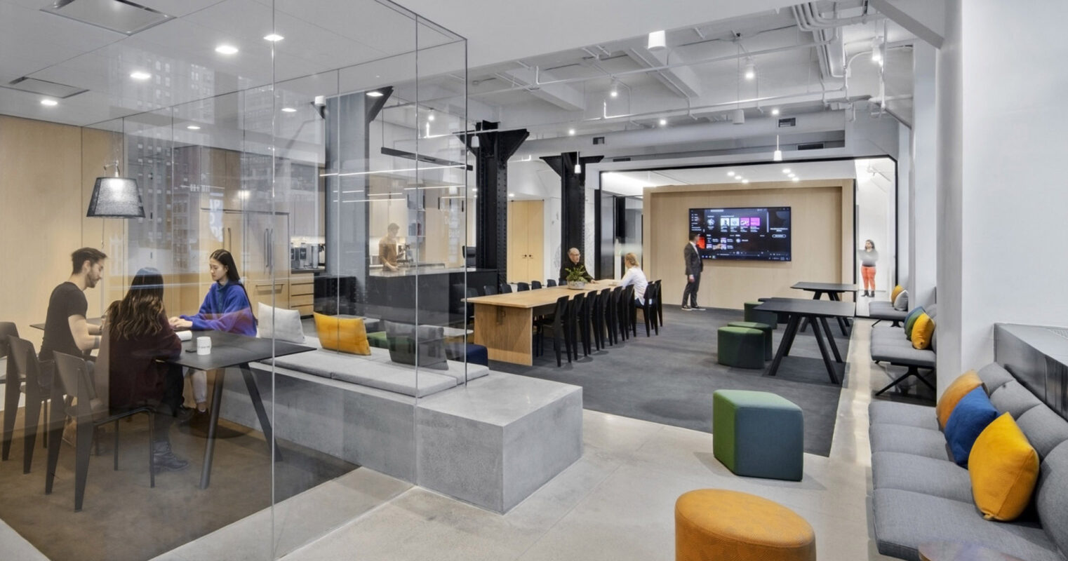 Modern office space showcasing a glass-walled meeting room, communal wooden worktable with pendant lighting, and casual lounge area with colorful, geometric furniture. Exposed ceiling and ductwork emphasize an industrial chic aesthetic contrasted with warm lighting for an inviting ambiance.