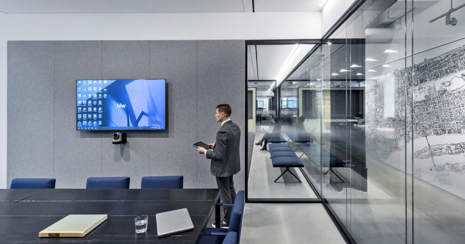 Modern office meeting room featuring sleek, glass walls, providing transparency and fostering a collaborative environment. The space is accentuated with a plush grey acoustic panel wall with an integrated flat-screen display, sophisticated furnishings, and innovative, detailed architectural drawings displayed as artistic elements. A professional stands, actively engaging with digital content.