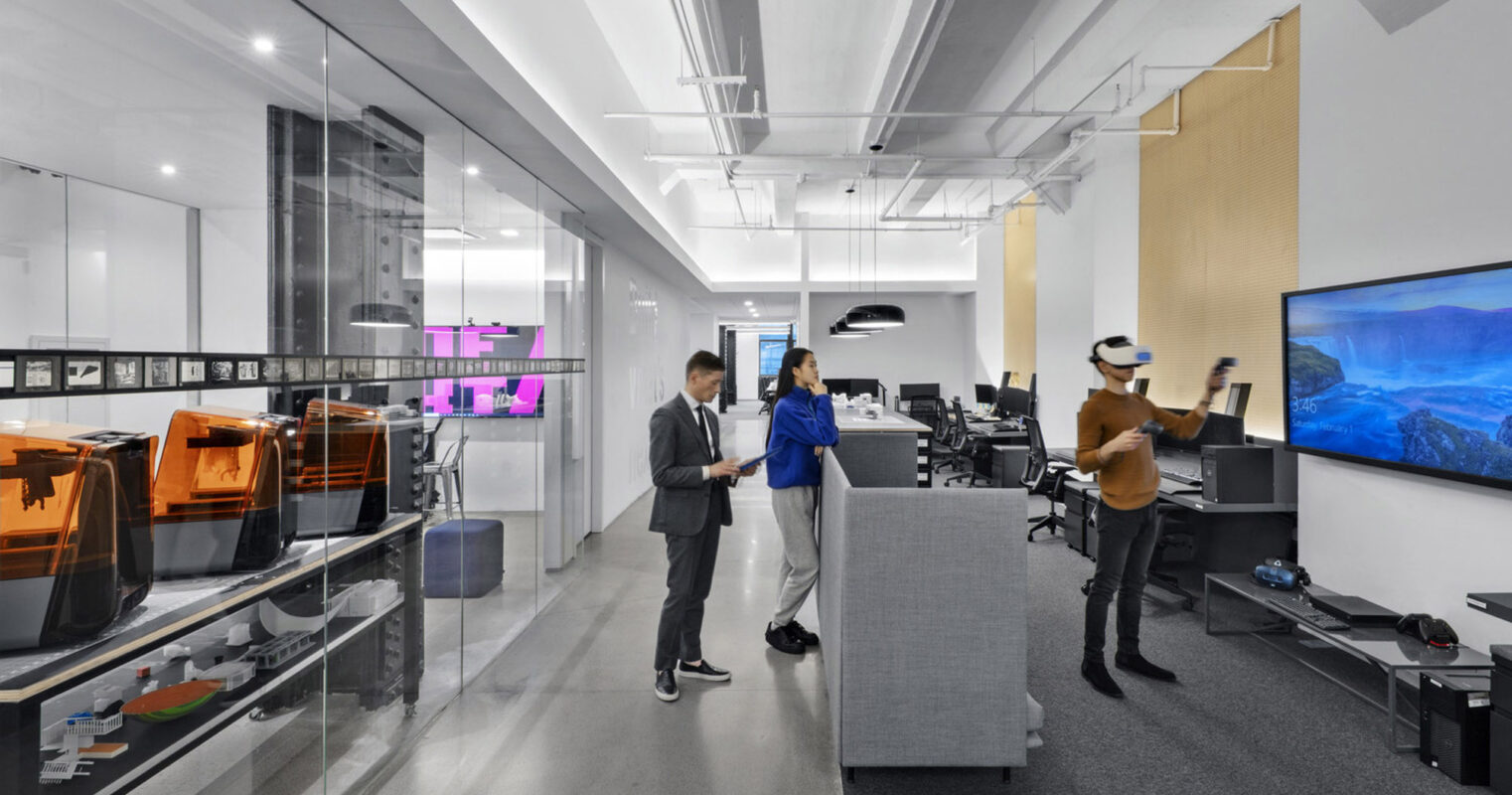 Modern office interior featuring vibrant orange 3D printers on the left, two professionals collaborating in the center, and a large blue-toned digital screen on the right wall, all under a neutral color palette with strategic pops of color.