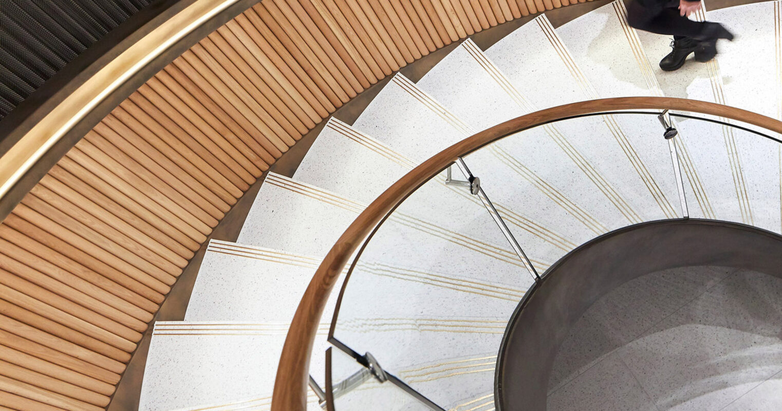 Elegant spiral staircase with warm wooden balustrades, contrasting the cool, grey tones of stone steps. A woman interacts with the fluid design, highlighting the staircase’s function and seamless integration within the space.