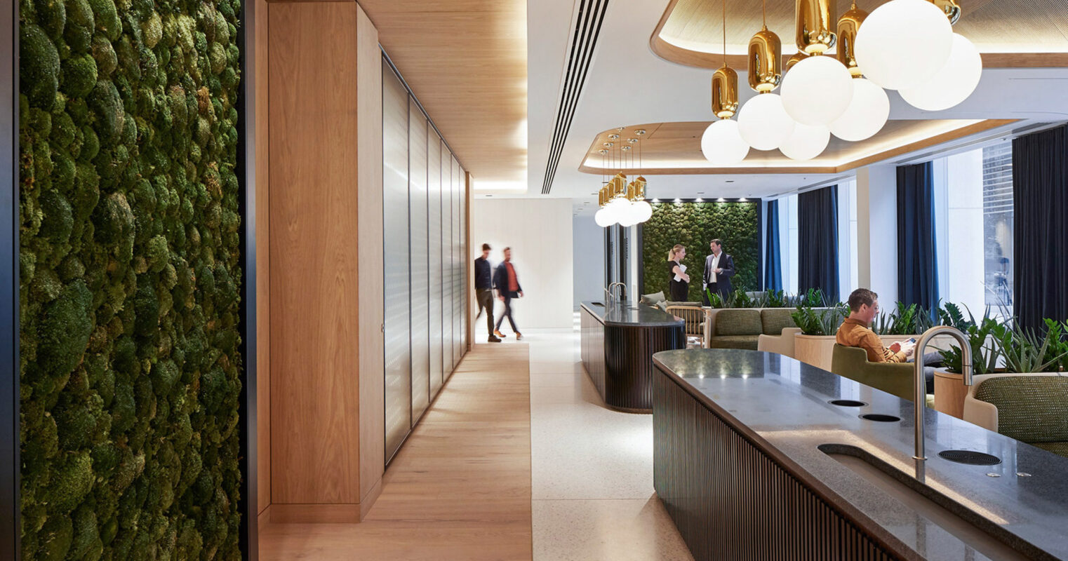 Modern office lobby with moss wall feature, wood paneling, and sleek reception desk. Golden pendant lights contrast with white spherical lamps above tailored seating areas.