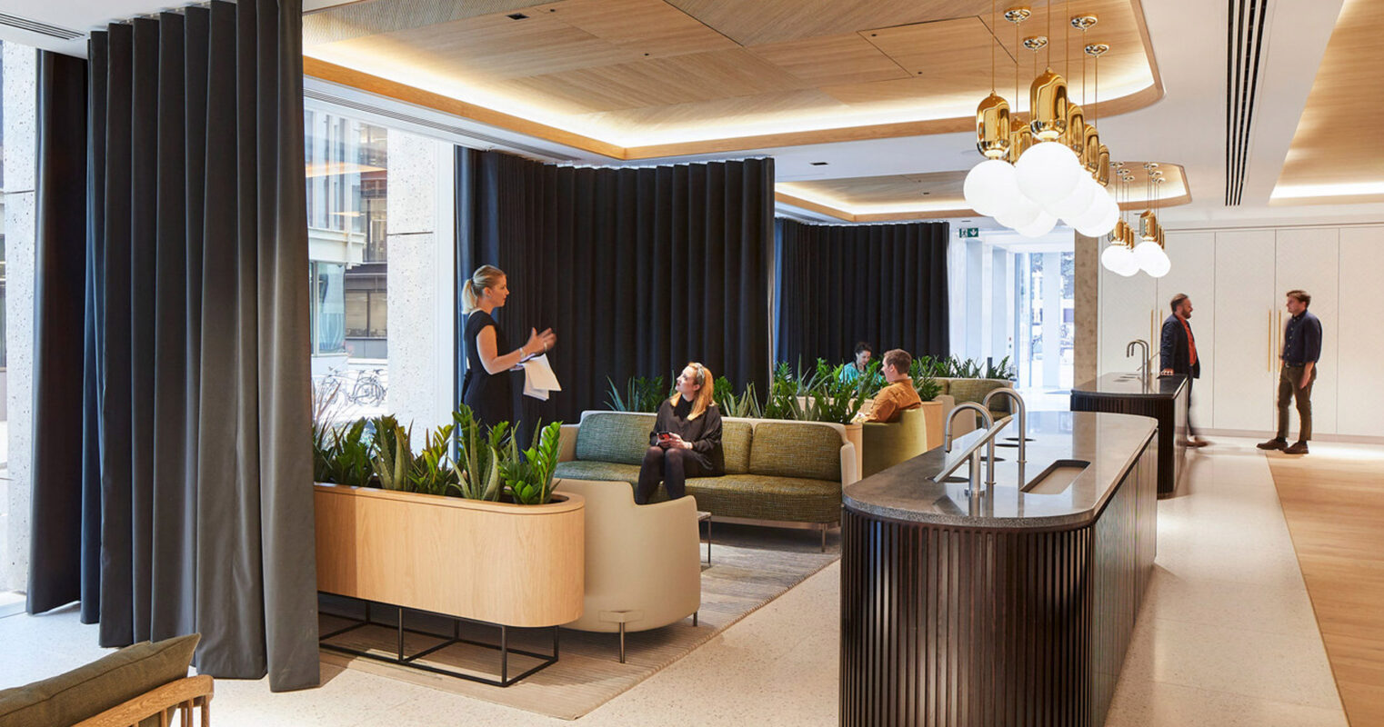 Modern office lobby with a warm, welcoming atmosphere, featuring curved wooden reception desk, elegant suspended light fixtures, and vertical drapery creating private nooks. Rich plant arrangements add a natural touch, while people converse and work, emphasizing the space's functional design.