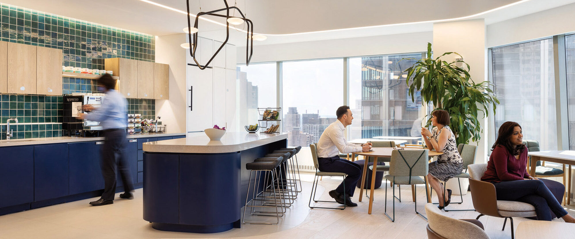 Modern office break room with exposed ceiling, featuring a central island with navy blue cabinetry and bar seating, surrounded by a wood-accent kitchen area, pendant lighting, and employees engaged in casual conversation.