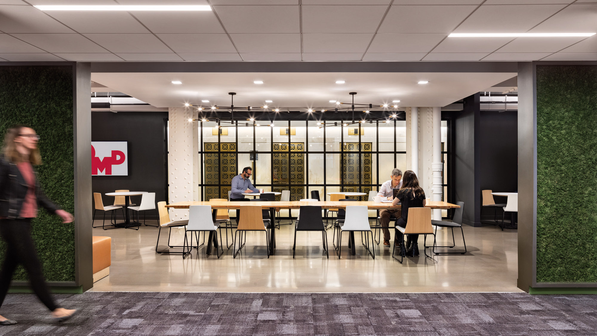 Modern office breakroom featuring polished concrete floors, light wood tables, and white Eames-style chairs under ambient lighting, with individuals collaborating casually. Exposed brick walls and a bold company logo accentuate the industrial-chic aesthetic of the space.