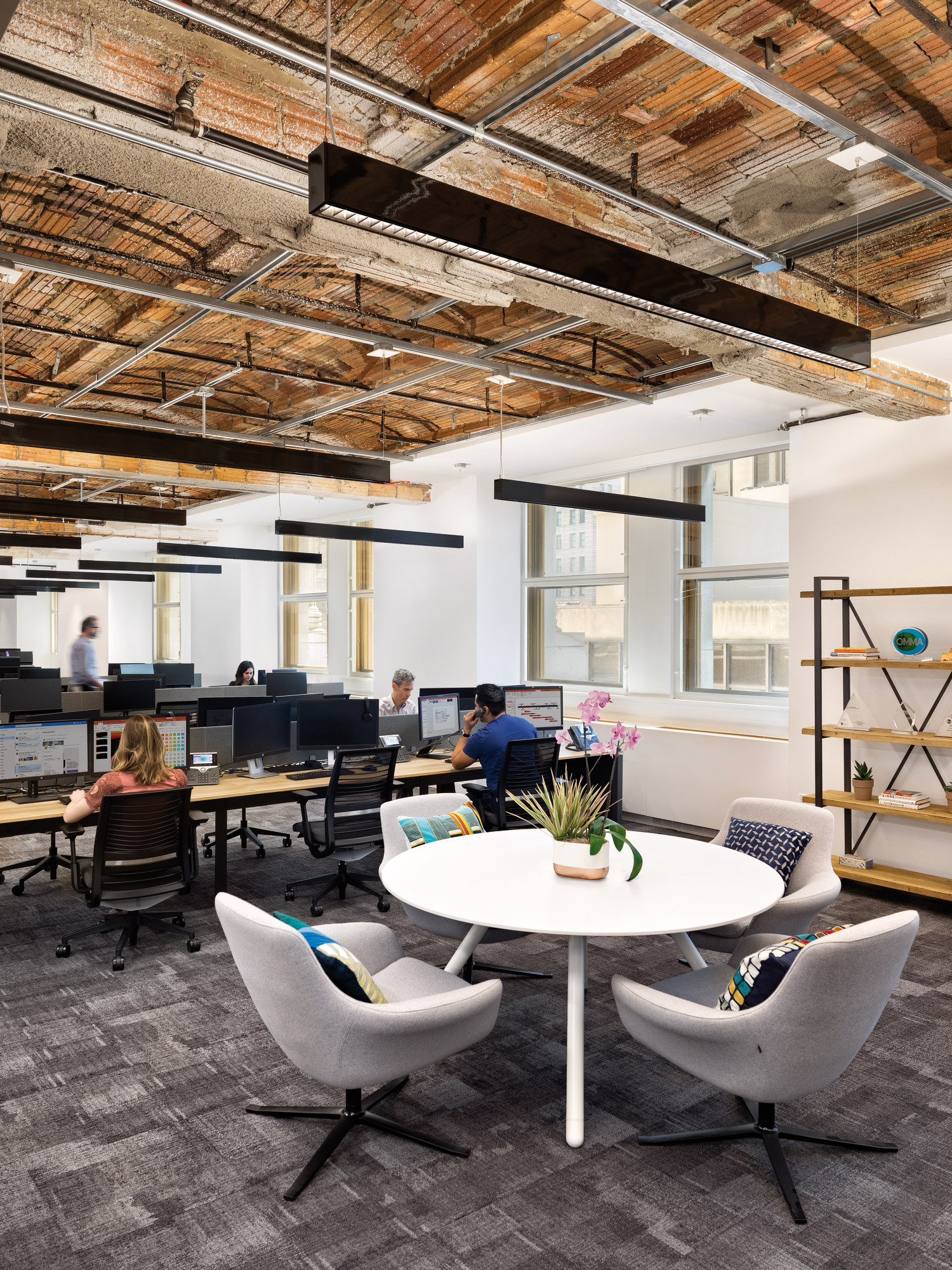 Modern open-plan office space featuring exposed wooden ceiling beams, natural daylight, ergonomic furniture with plush gray seating, and a minimalist white round table centering a collaborative area. Ambient lighting enhances the warm, productive atmosphere amidst neutral color tones and green plant accents.
