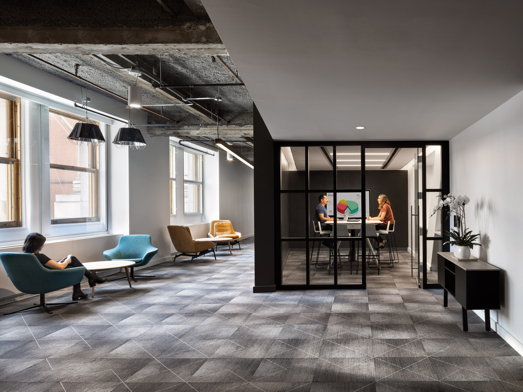 Spacious modern office lobby featuring geometric patterned carpet, cozy seating areas with colorful mid-century chairs, a sleek black-framed glass meeting room, and industrial-style exposed ceiling beams. Natural light streams in through large windows, complementing the warm ambient lighting.