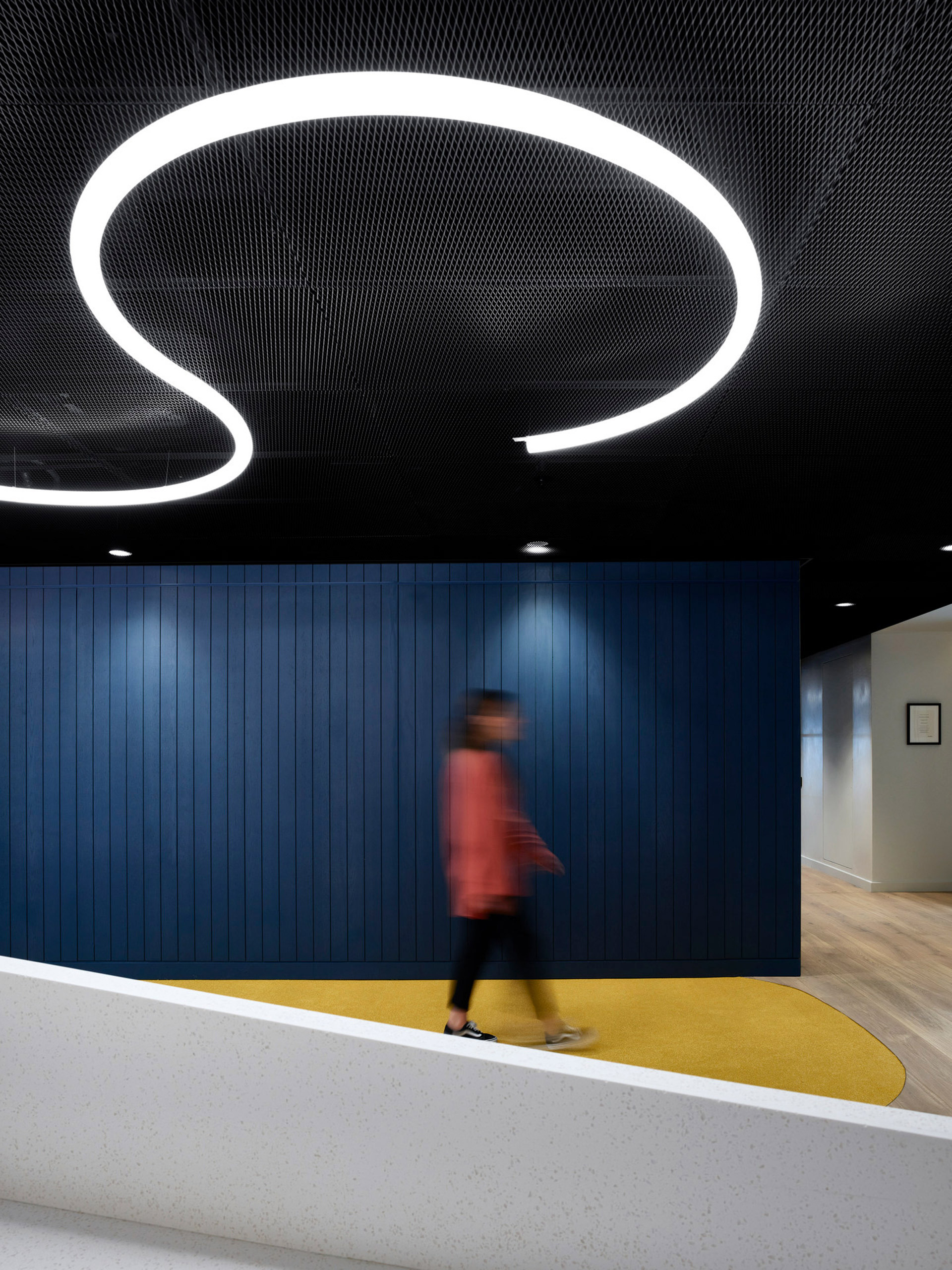 Person walks past a striking reception area characterized by a bold overhead spiral lighting fixture, casting a warm glow on the textured dark ceiling. Below, a curving yellow carpet complements the clean lines of the blue paneled walls and sleek white reception desk.