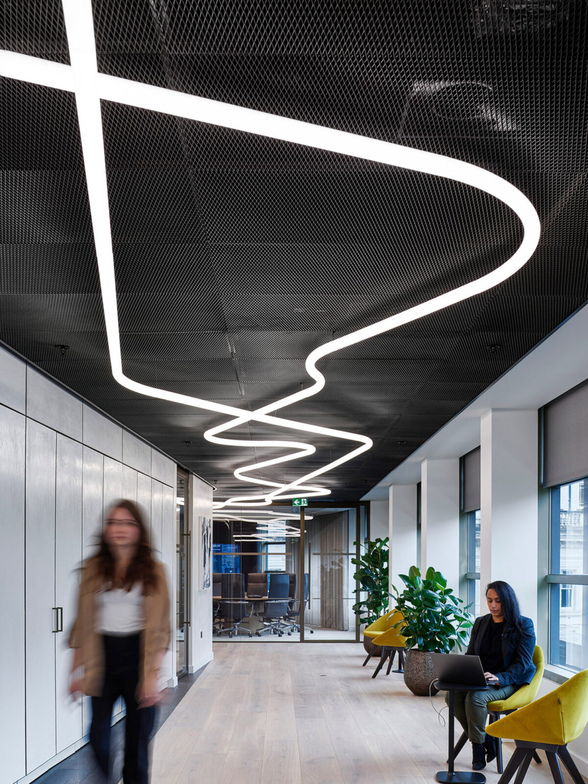Modern office lobby with dynamic LED lighting gracefully snaking across a black mesh ceiling. The scene includes wood flooring, sleek dark wall panels, and vibrant yellow accent chairs. Individuals are engaging in casual work activities in a contemporary, energized environment.