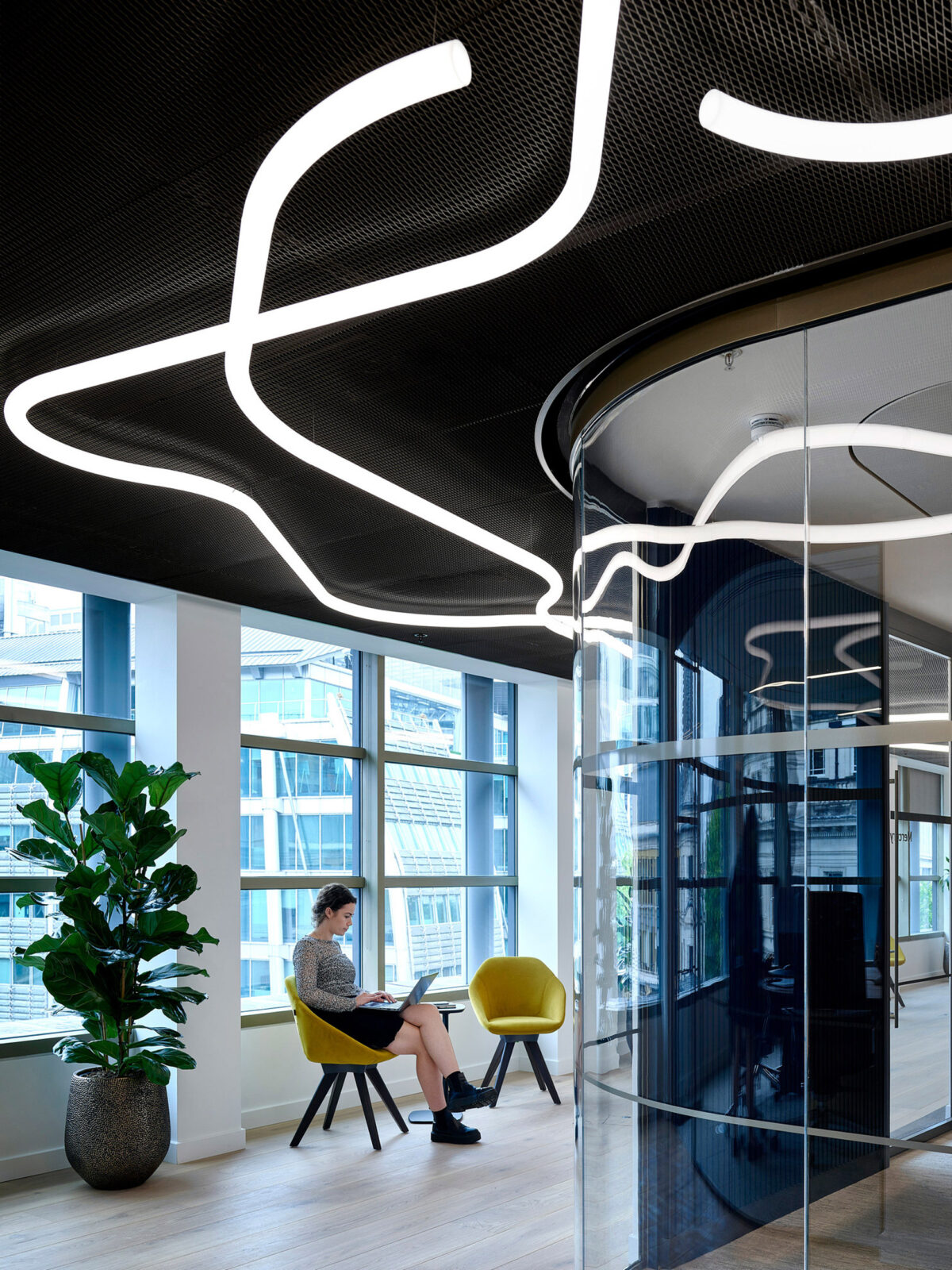 Contemporary office space featuring a striking overhead serpentine lighting design, contrasted against a black ceiling. The sleek, circular glass meeting room adds transparency, complemented by a vibrant yellow chair and lush greenery, enhancing the visual aesthetic and collaborative environment.