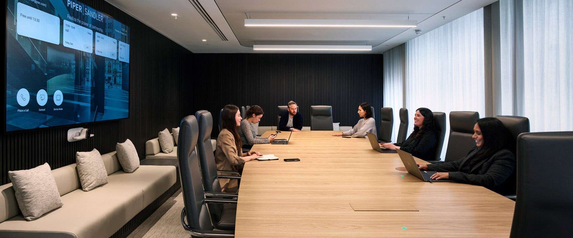 Modern boardroom with a sleek, wooden conference table, surrounded by ergonomic chairs. A large digital screen displays a presentation, while natural light filters through the windows, complementing the room's recessed and strip lighting.