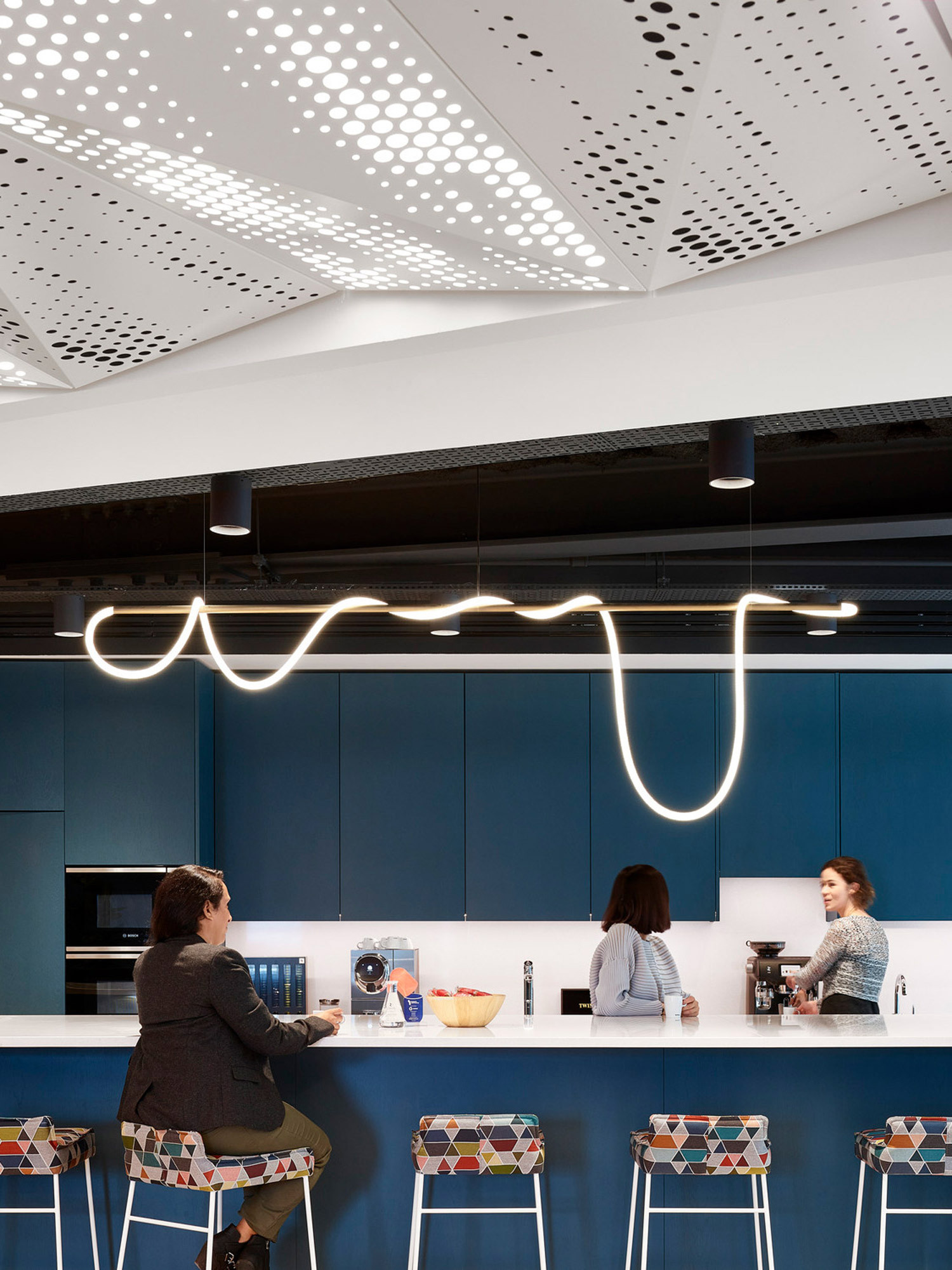 Modern kitchen workspace featuring cobalt blue cabinetry, whimsical overhead LED lighting resembling a squiggled line, and geometrically patterned bar stools. Perforated ceiling panels add texture and acoustic functionality above a conversational seating area.