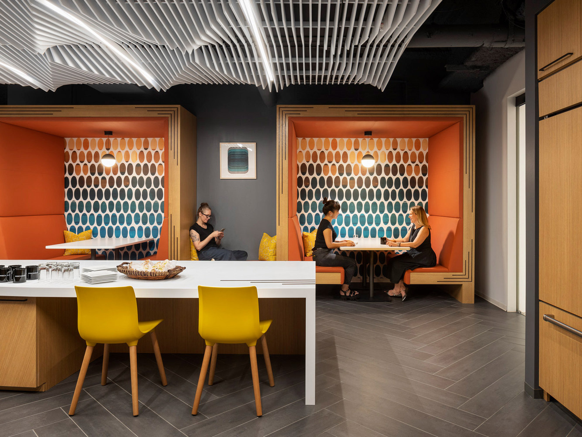 Bright, modern office breakroom featuring undulating ceiling panels, vibrant orange booths, honeycomb-patterned dividers, and bold yellow chairs around a minimalist white table. Natural wood accents complement the space's dynamic and inviting atmosphere.