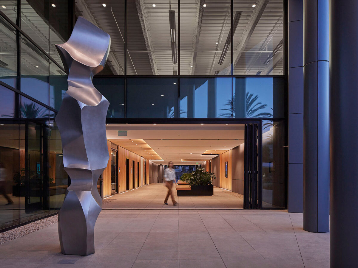 Elegant commercial building entrance featuring a towering abstract sculpture, accentuating the space with its reflective metallic surface under warm, recessed LED lighting. The clean lines of the architecture are complemented by tropical foliage, enhancing the inviting walkway.