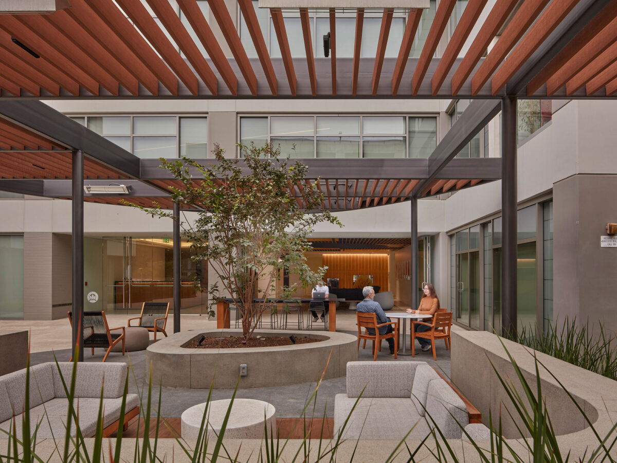 Elegant courtyard with a modern design, featuring a central tree planter, surrounded by wooden bench seating. Overhead, parallel wooden slats form a pergola, casting linear shadows. Through floor-to-ceiling windows, the interior's warm lighting contrasts the serene outdoor space, populated with contemporary furnishings and conversing individuals.
