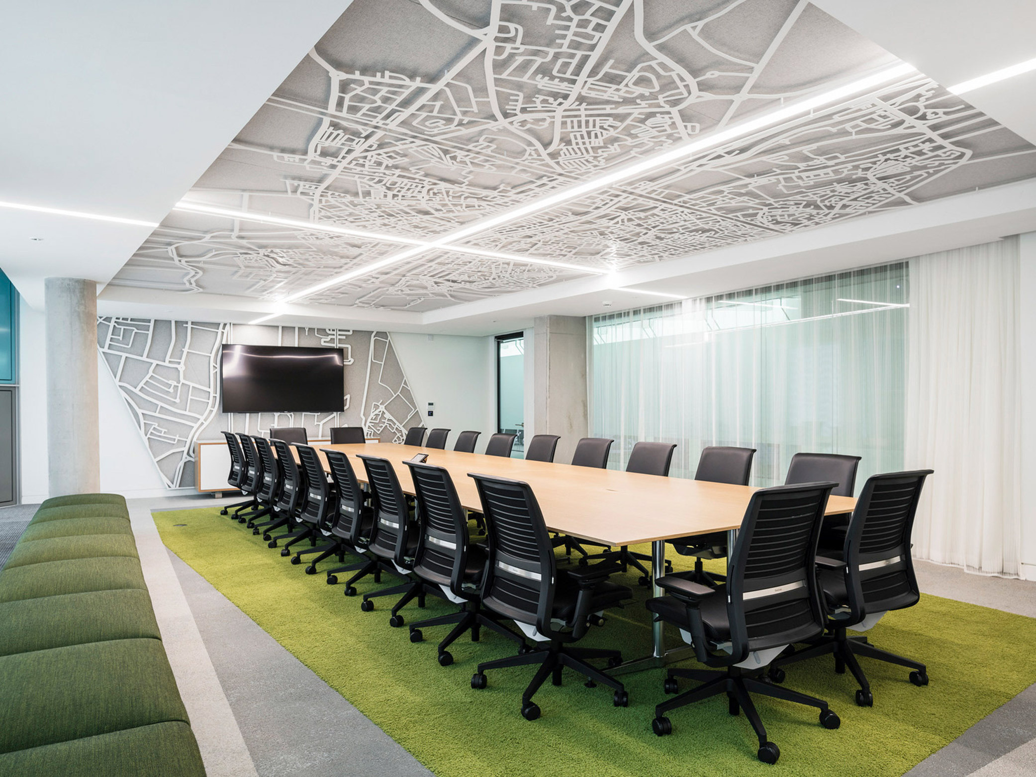 Modern conference room featuring an elongated wooden table surrounded by black office chairs, with a vibrant green carpet mimicking grass. An artistic ceiling panel displays a white abstract city map design, complemented by recessed lighting. Large screen and white drapes adorn the adjacent wall.