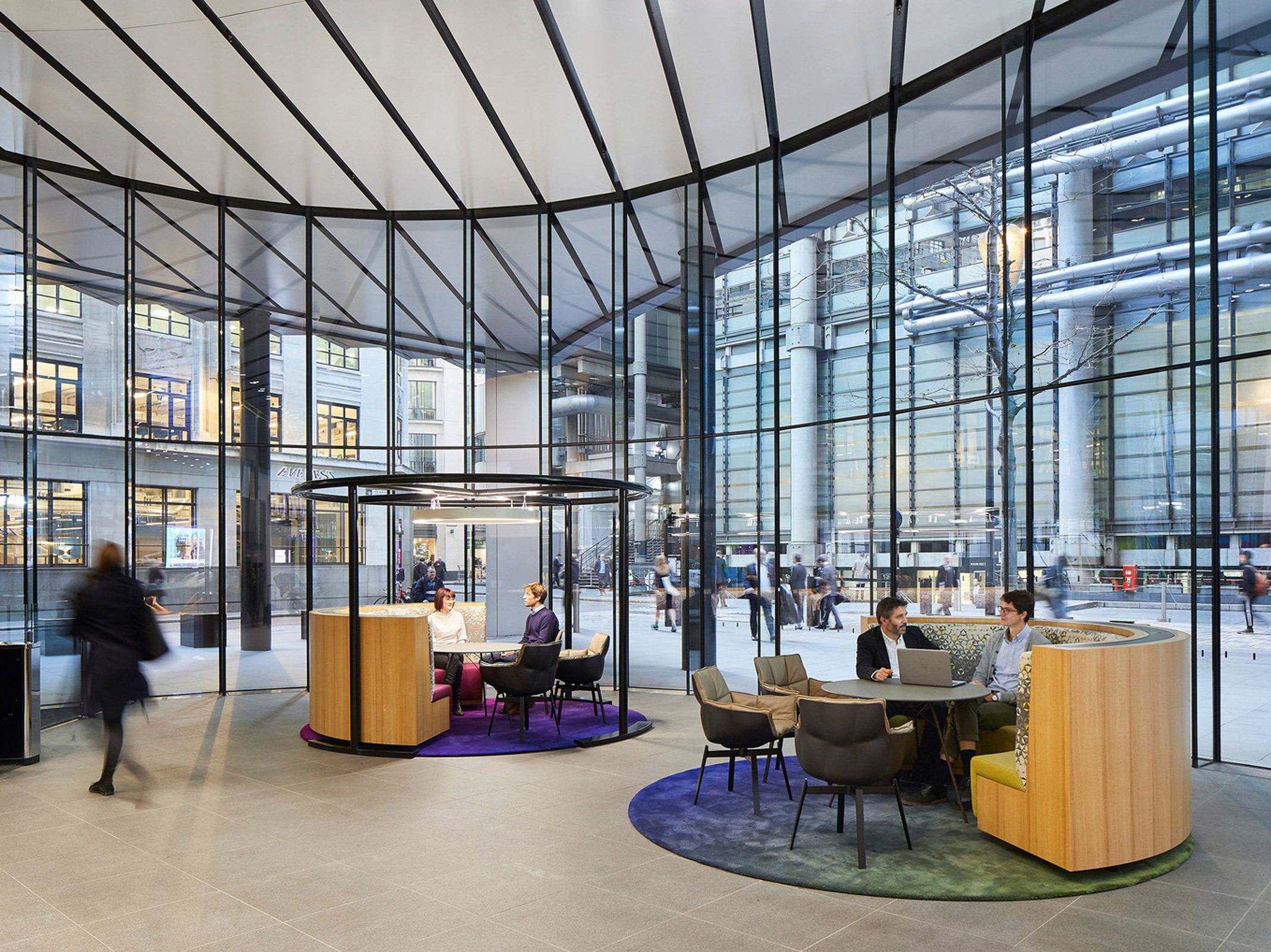 Open-plan office lobby with floor-to-ceiling glass walls. Central seating pod with integrated technology, flanked by casual chairs and felt dividers. Dynamic contrast between bustling exterior and focused collaborative spaces within.