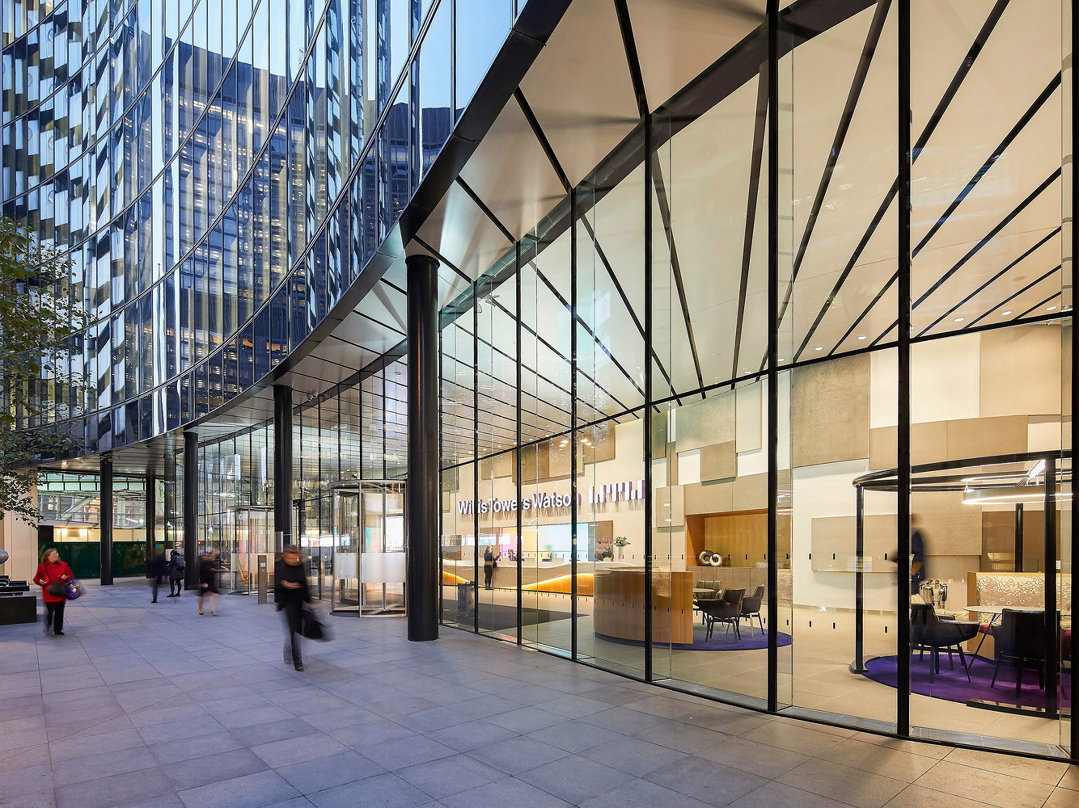 Modern commercial building interior visible through floor-to-ceiling glass facade; sleek design featuring natural stone walls, minimalist furniture with pops of purple accents, and warm lighting enhancing an inviting, open-plan layout.