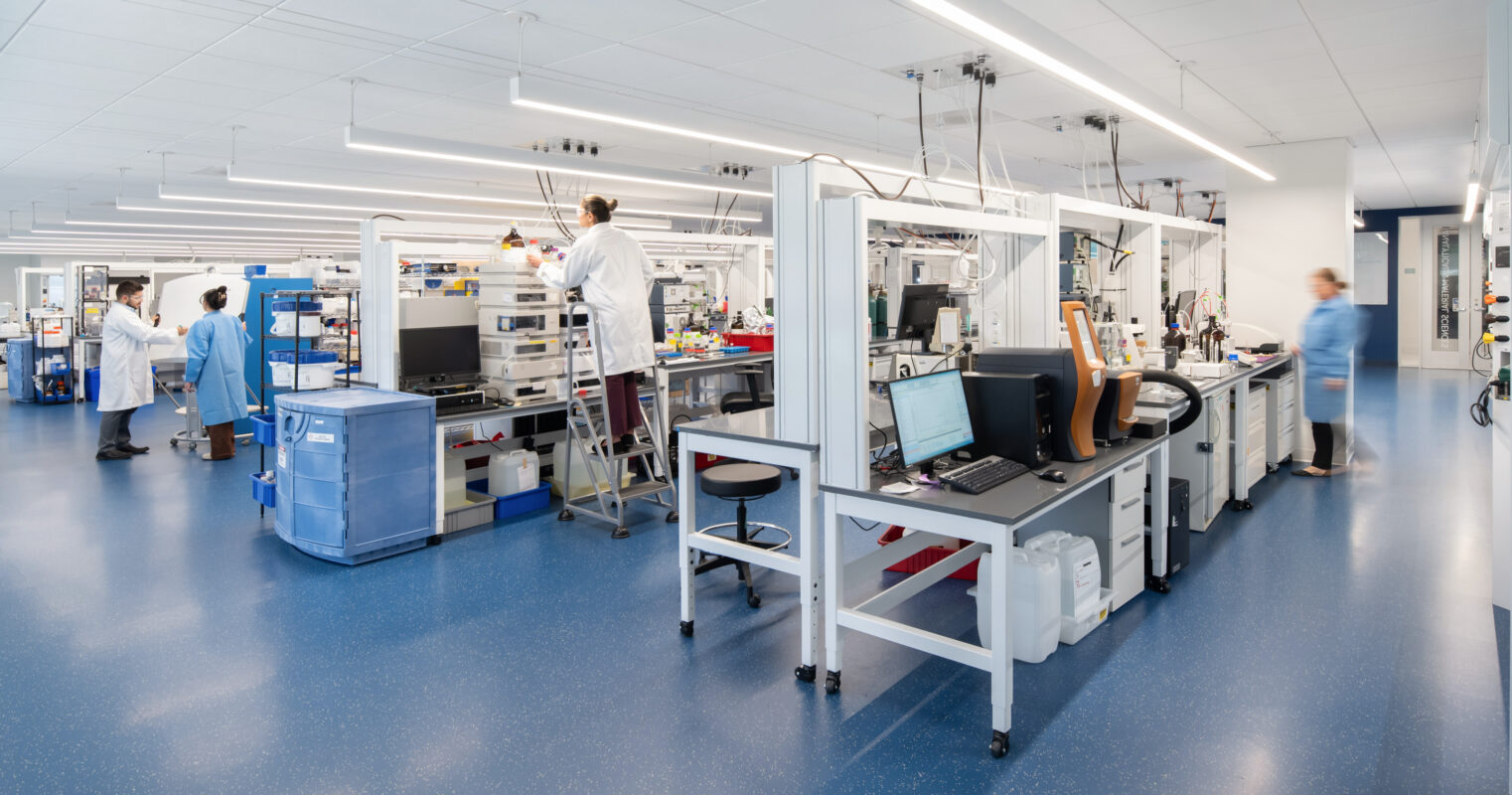 Modern laboratory space featuring streamlined workstations equipped with advanced technology. The open layout promotes collaboration, allowing researchers to interact freely. Bright overhead lighting and smooth, blue flooring enhance visibility and cleanliness, while laboratory equipment is neatly organized to create an efficient workflow. People in lab coats engage in various tasks.