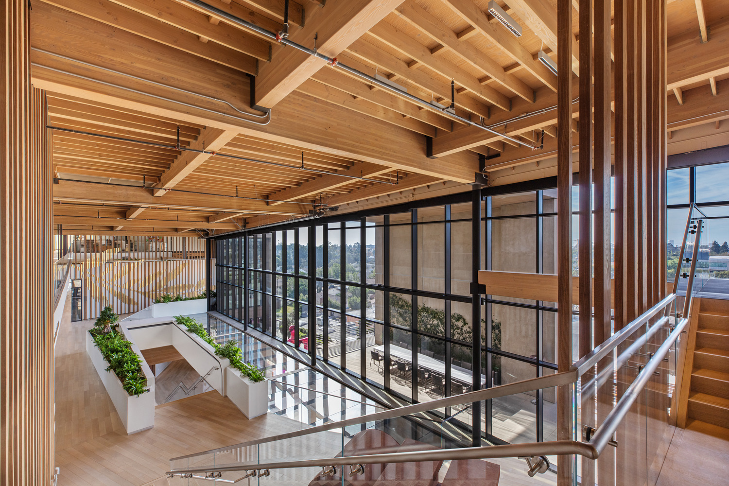 Modern office interior highlighting exposed wooden beams and slatted ceiling design, complemented by glass railing, open-plan staircases, and indoor greenery, creating a biophilic, industrial aesthetic with natural light.