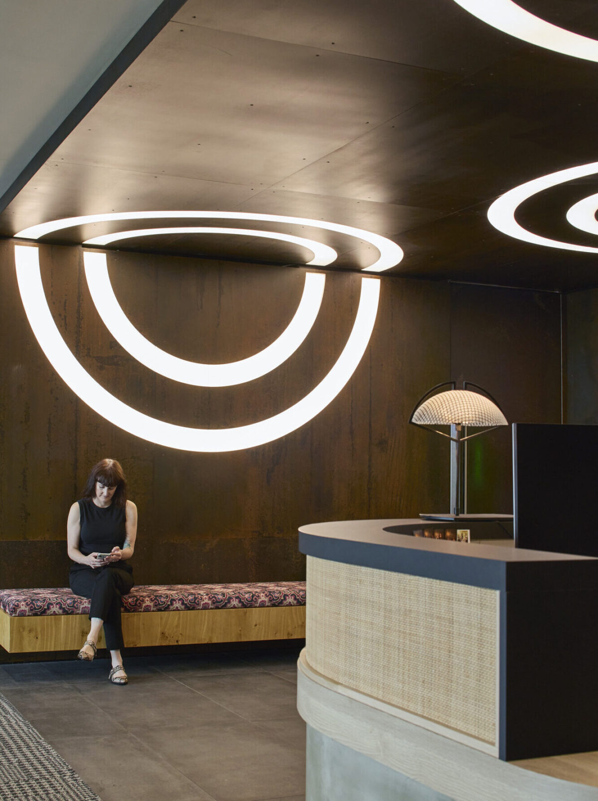 A contemporary reception area features a sleek, rounded wooden bench with patterned upholstery, illuminated by striking circular light fixtures. A woman sits quietly, contrasting with the industrial metallic wall. The curved reception desk, made of textured wood, complements the modern aesthetic, enhancing the inviting atmosphere.