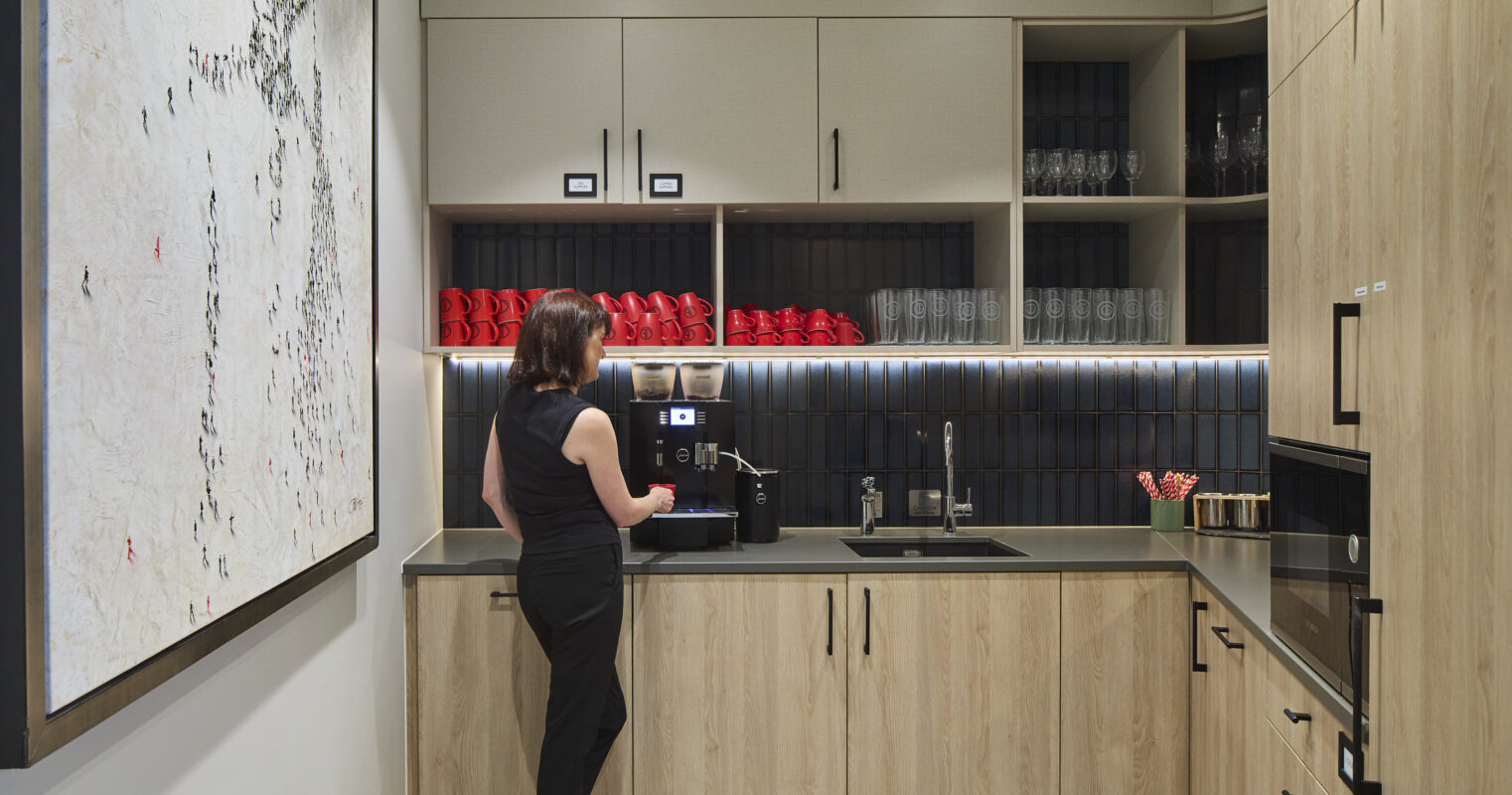 A modern kitchen features sleek wooden cabinetry with contrasting black countertops. A large art piece complements the minimalist aesthetic. A woman stands near a coffee machine, highlighting the functional yet stylish design. The combination of textures and colors creates a warm, inviting atmosphere ideal for relaxation and socializing.