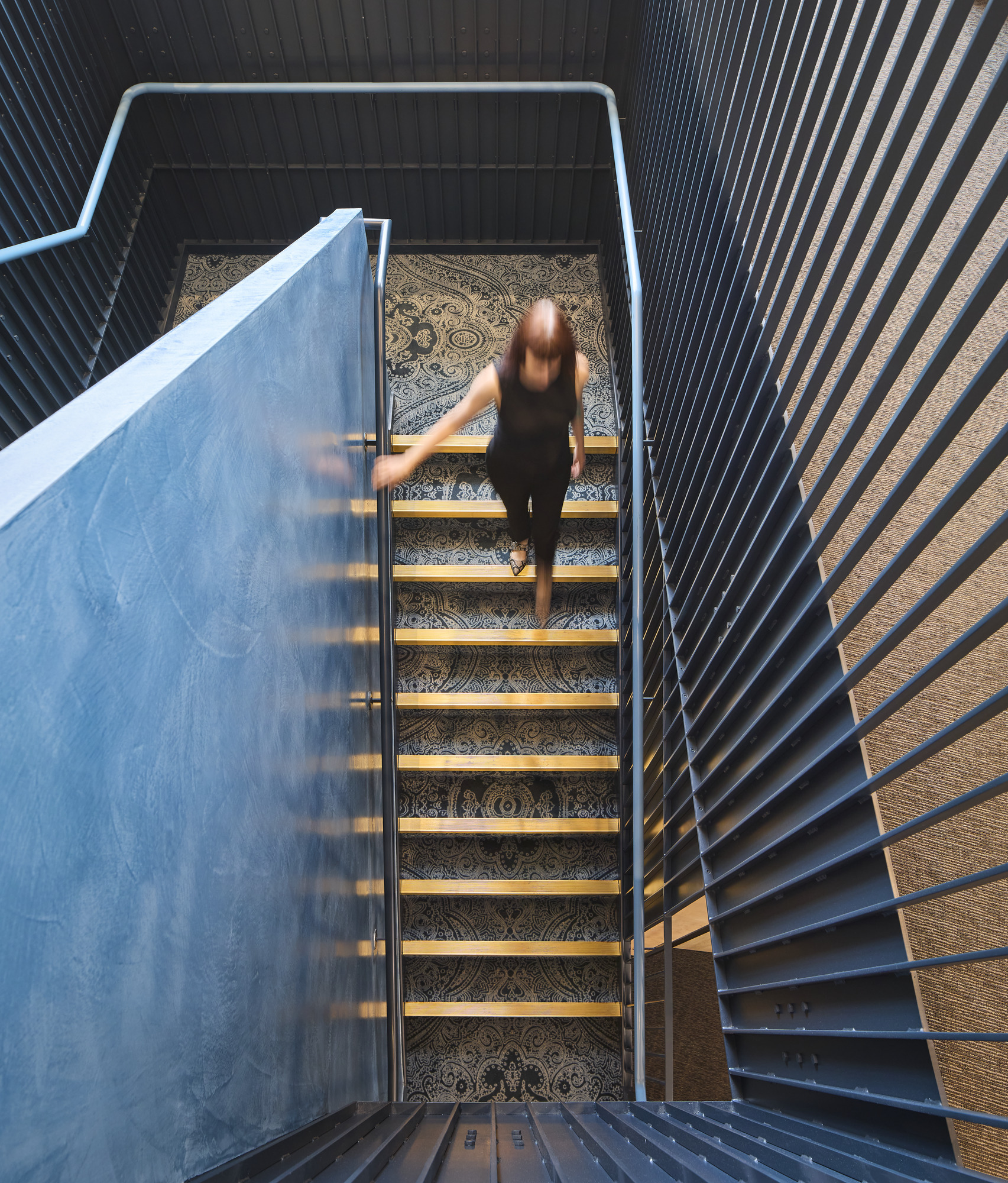 A person descends a staircase featuring a modern design with metal railings and intricate patterned carpet. The walls are lined with vertical wood slats, enhancing the contemporary aesthetic. Soft lighting creates a welcoming atmosphere, highlighting the interplay between textures and materials in the space.