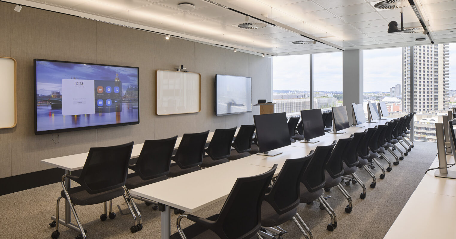 Modern conference room featuring sleek, ergonomic chairs arranged at long, white tables. Large windows offer expansive city views, while integrated technology includes multiple screens mounted on a muted beige wall. The design emphasizes functionality and collaboration, complemented by a neutral color palette and ample overhead lighting.