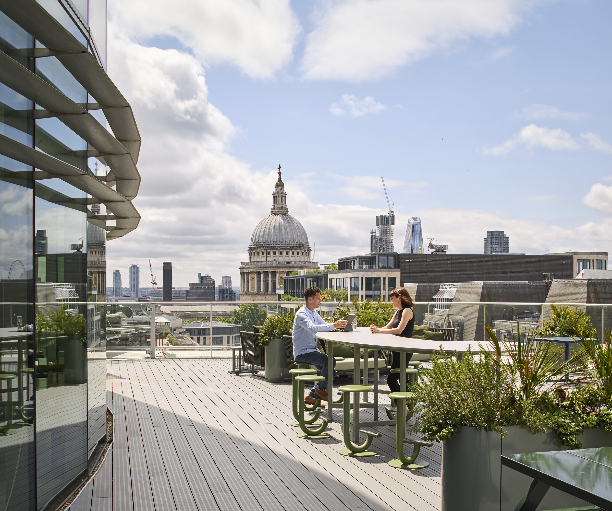 A vibrant rooftop terrace features modern seating arrangements surrounded by lush greenery. Two individuals engage in conversation at a sleek table, with a backdrop showcasing London’s iconic St. Paul's Cathedral and contemporary skyscrapers, blending urban life with outdoor relaxation in a stylish setting.