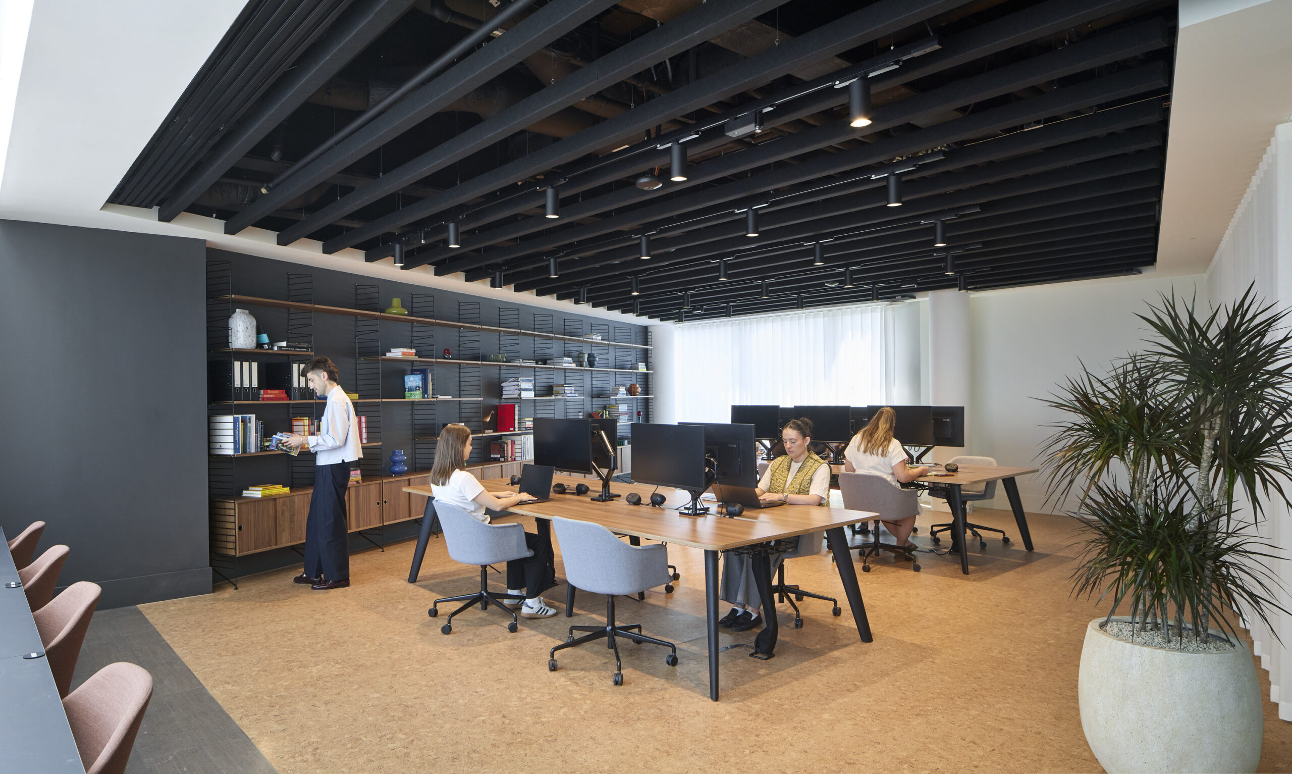 A modern open-plan office featuring modular desks arranged for collaborative work. Natural light floods the space, highlighting the warm cork flooring. A dark-beamed ceiling contrasts with light walls, while stylish storage units and potted plants add to the contemporary aesthetic, promoting productivity and creativity.