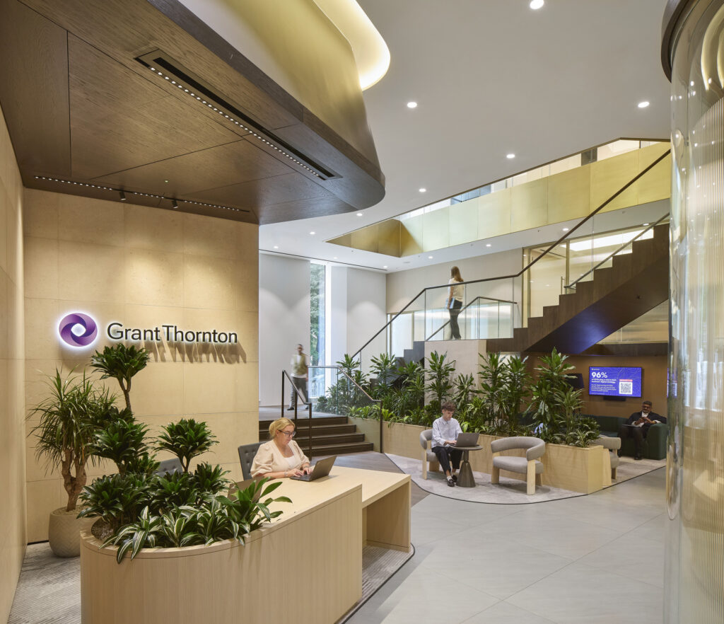 The spacious reception area of Grant Thornton features sleek wood paneling and modern furnishings. Lush greenery adds a touch of nature, while sophisticated lighting casts a warm glow. A prominent staircase leads to upper levels, enhancing visual interest and welcoming clients and guests into a refined workspace.